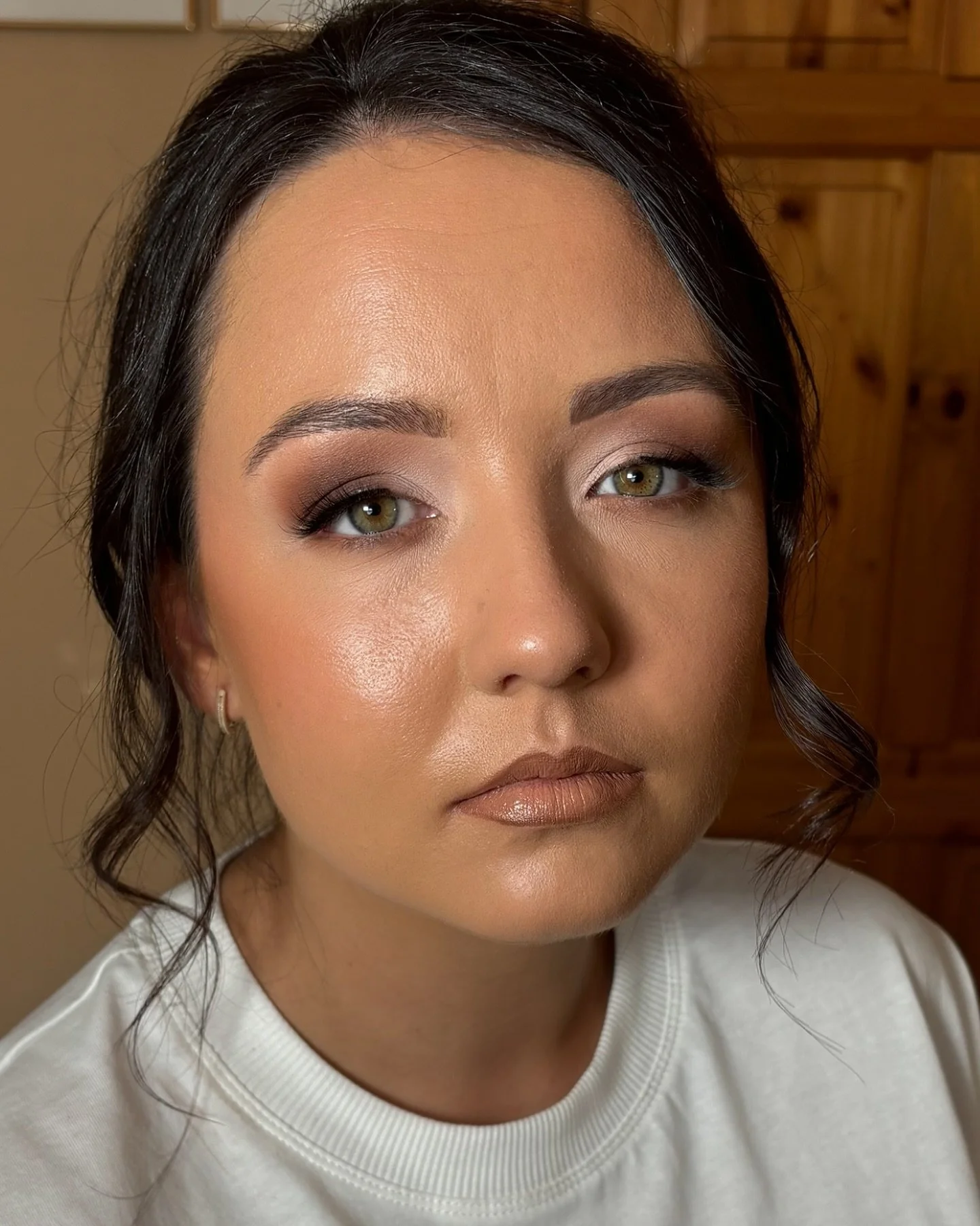 Close-up of a woman with styled dark hair, green eyes, and makeup including eyeshadow, eyeliner, mascara, and nude lipstick, wearing a white top, against a wooden background.