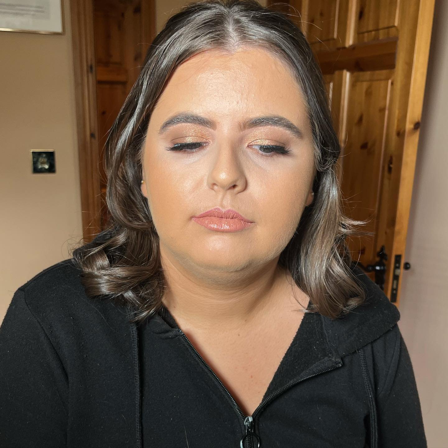 A woman with shoulder-length brown hair, styled in loose curls, wearing makeup with gold eyeshadow, nude lipstick, and a black zip-up top, standing indoors with a wooden door and beige walls in the background.
