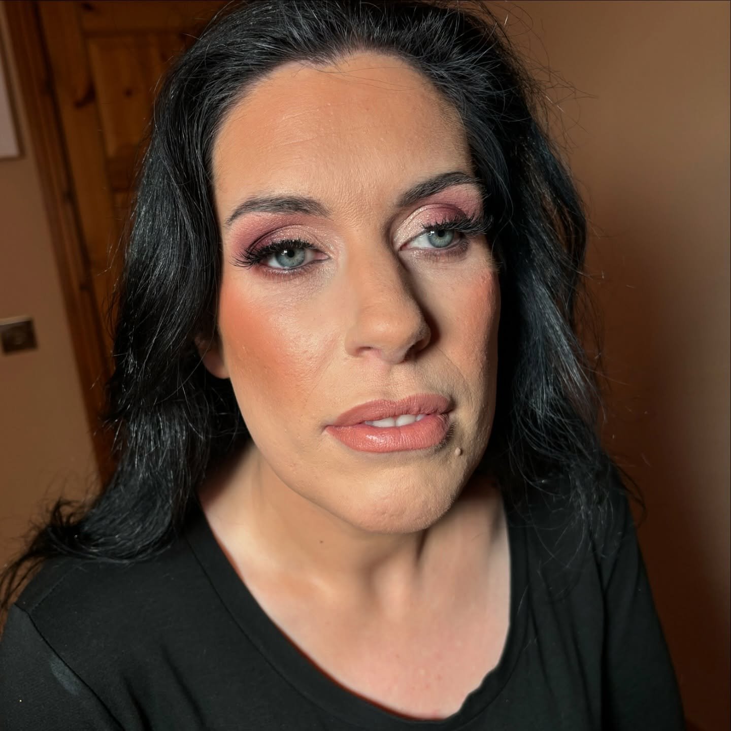 Close-up portrait of a woman with long black hair, blue eyes, and makeup, wearing a black top, standing indoors with a wooden door in the background.