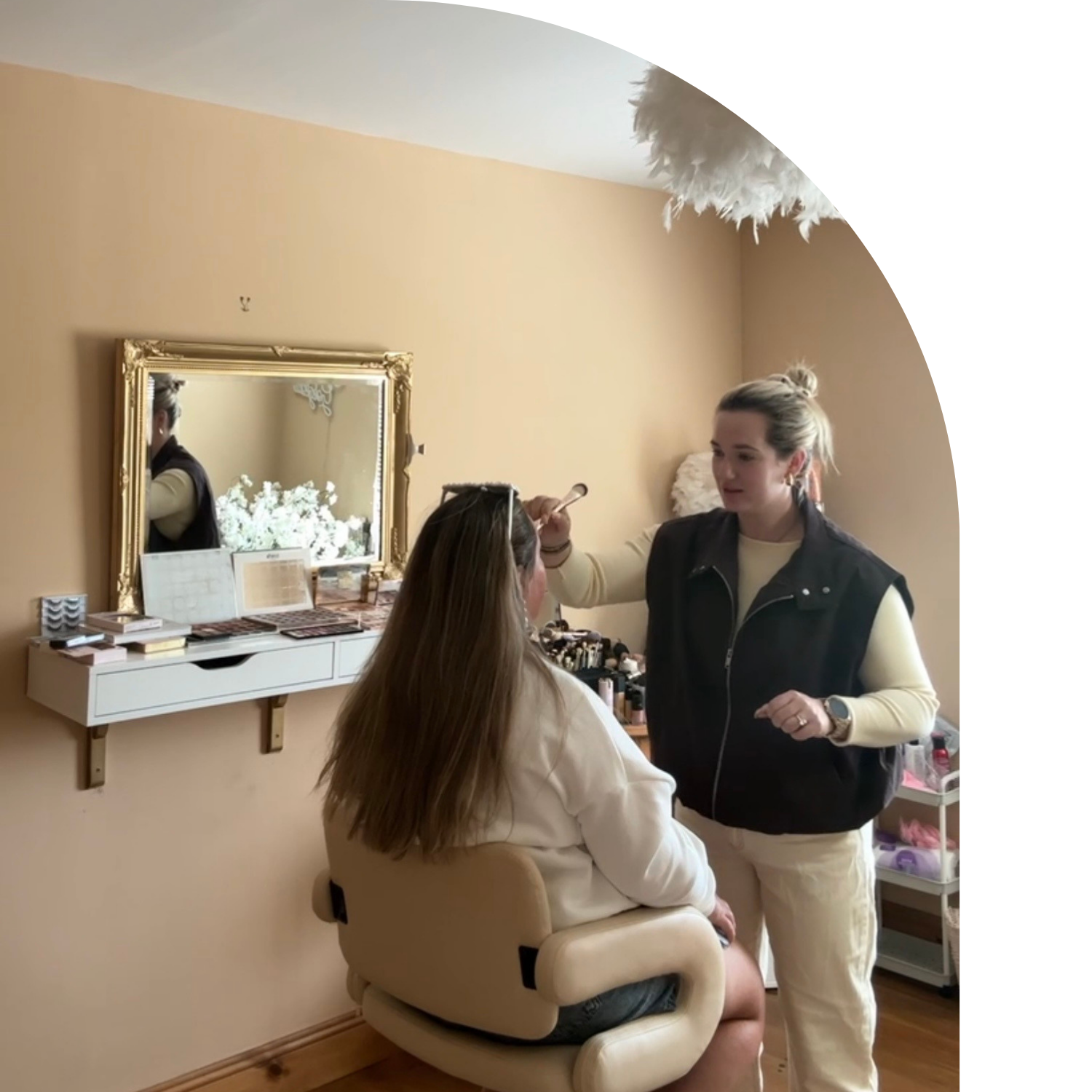 A woman with long brown hair seated in a beauty salon chair, getting her makeup done by a makeup artist with blonde hair tied in a bun. The salon has beige walls, a white shelf with makeup supplies, and a large gold-framed mirror reflecting part of the room.