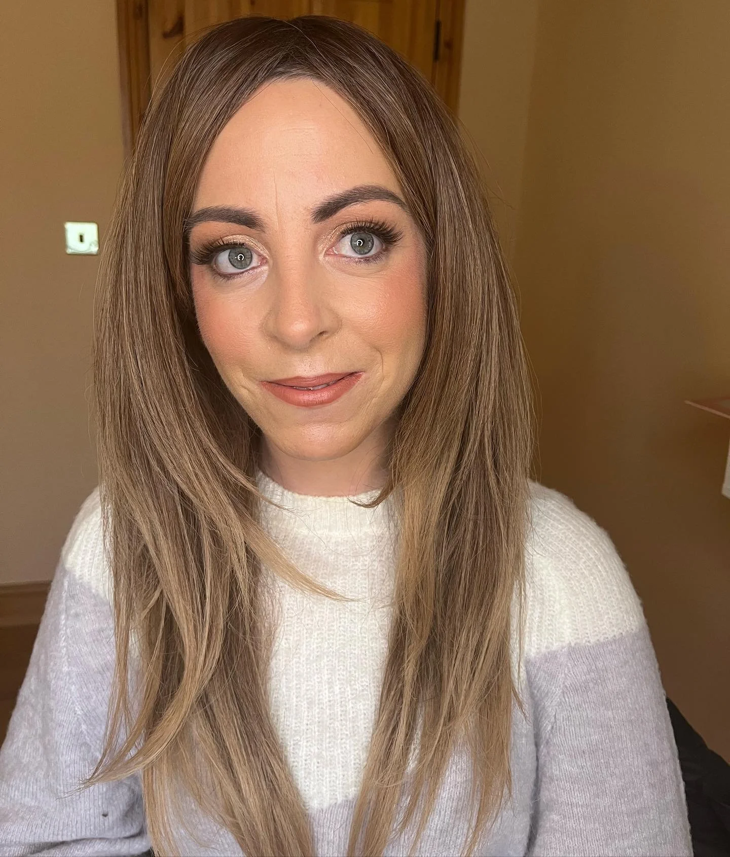 A young woman with long, light brown hair and blue eyes wearing a white sweater, smiling softly at the camera in an indoor setting with wooden background and yellow walls.