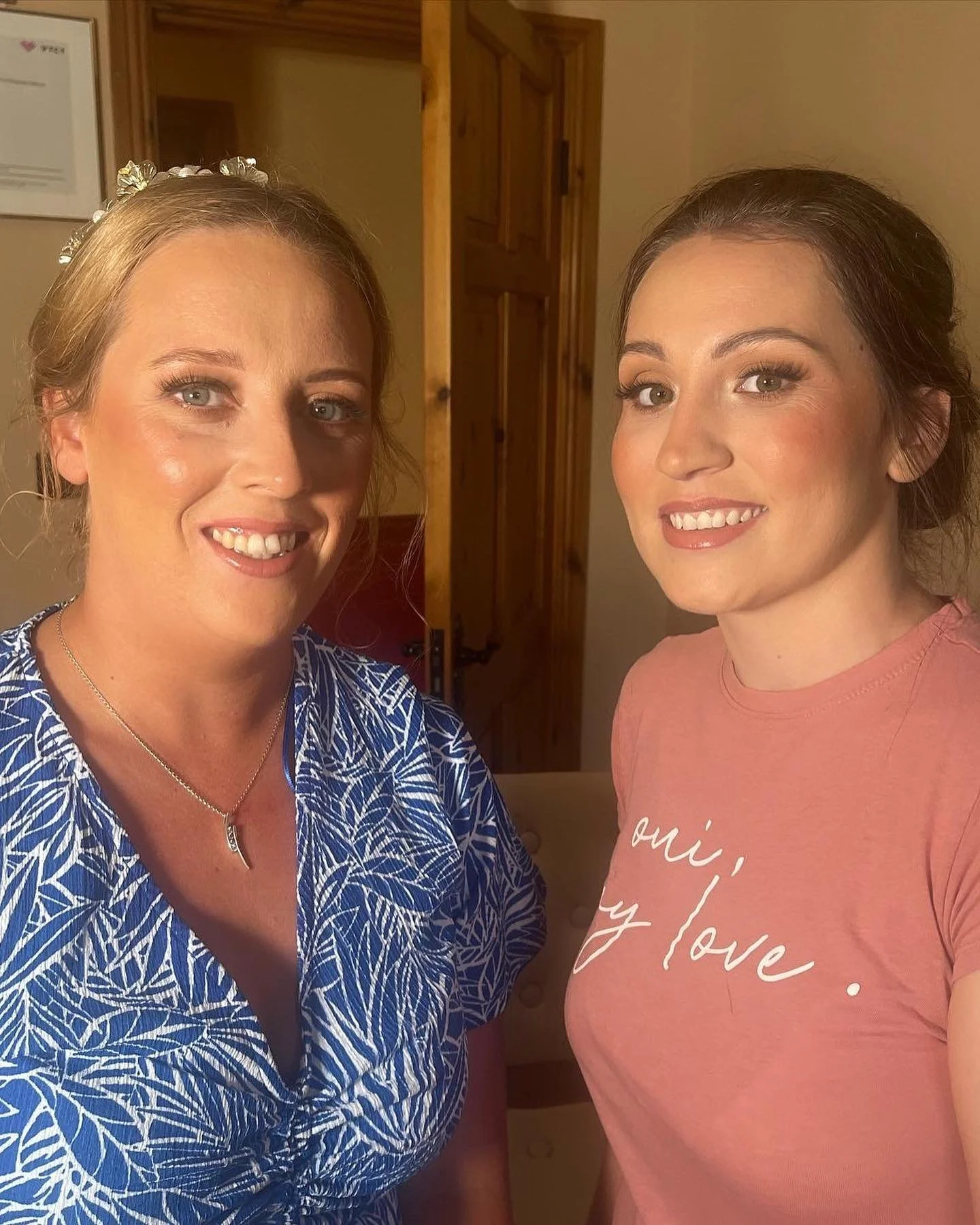Two women smiling and looking at the camera, standing indoors with wooden doors and beige walls in the background. One is wearing a blue patterned shirt with a silver necklace, and the other is wearing a pink shirt with white text.