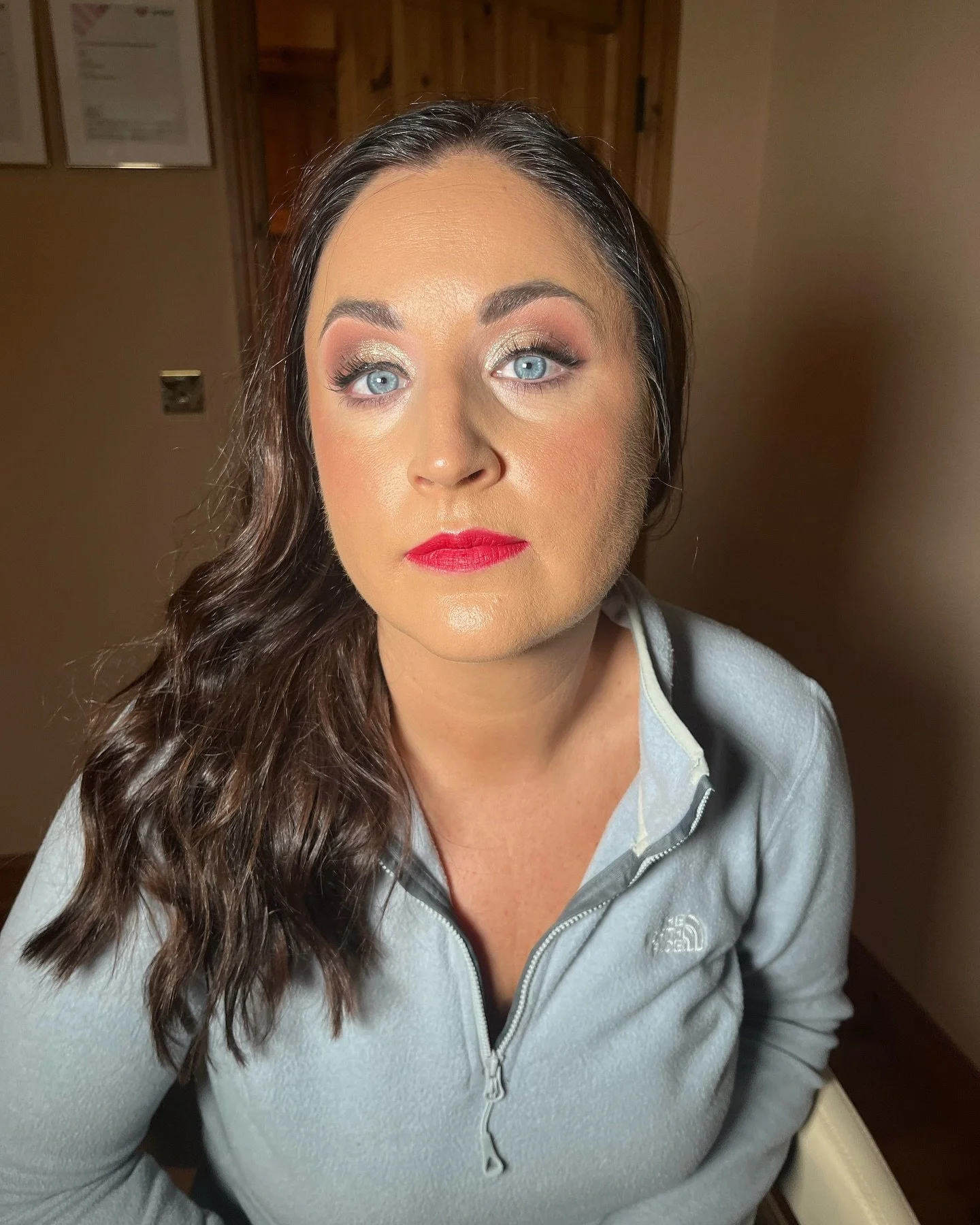 A woman with long, wavy brown hair, blue eyes, and makeup including gold eyeshadow and red lipstick, wearing a light gray zip-up jacket, sitting indoors.