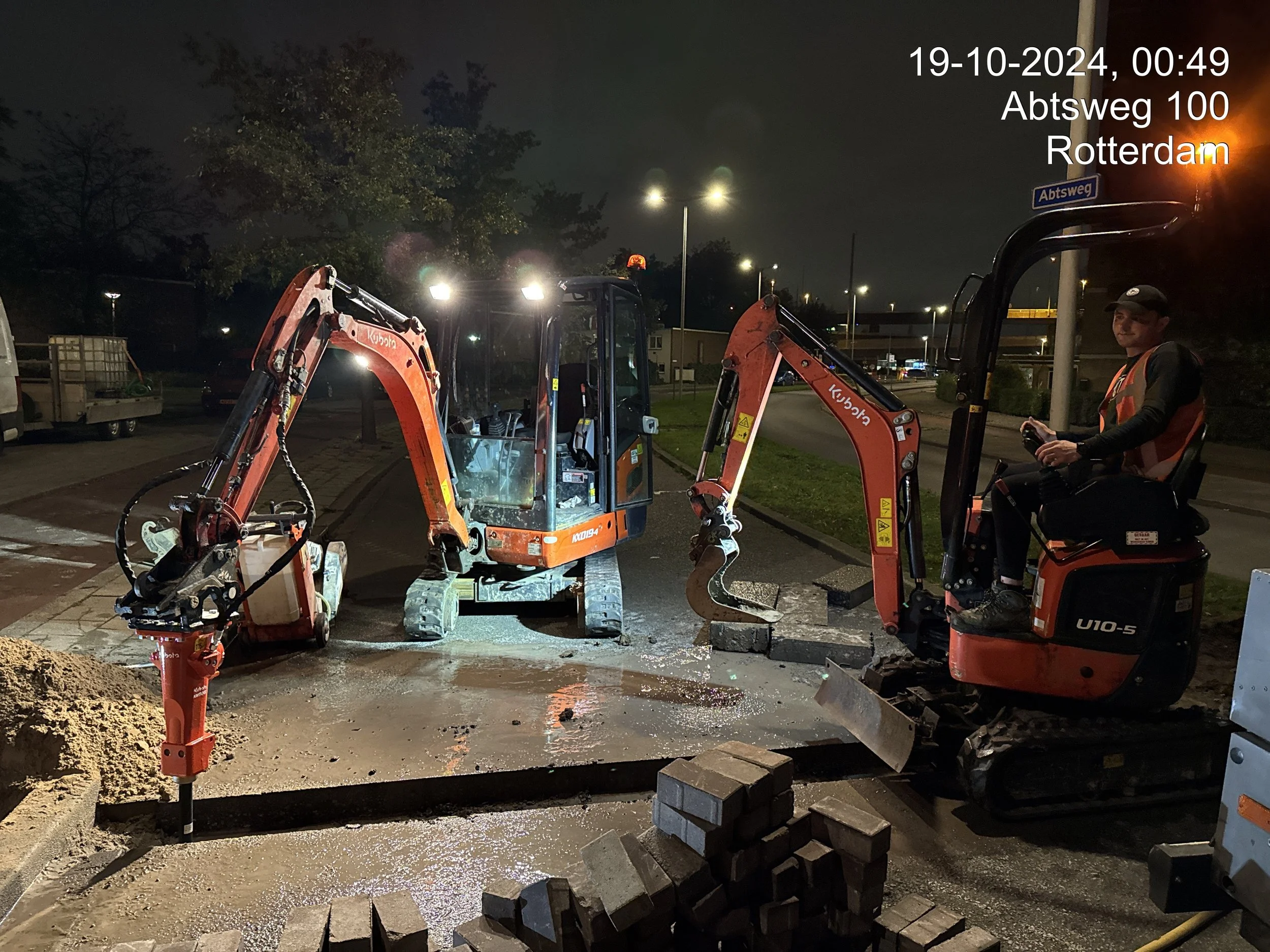 Nighttime scene of road construction in Rotterdam with construction equipment, a worker on a mini excavator, and streetlights illuminating the area.