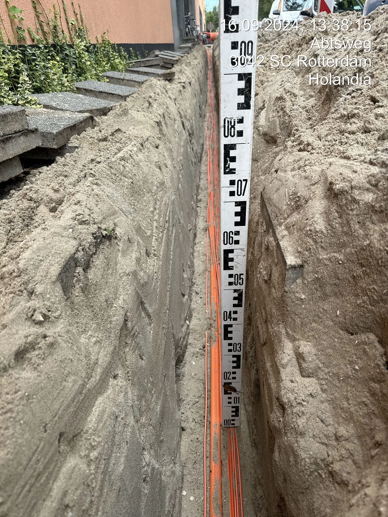 A construction site with an open trench and a measuring rod showing depth. On the left, there are stacked concrete slabs and some green plants. The background includes parked vehicles and a building. The image contains an overlay with date, time, and location details.