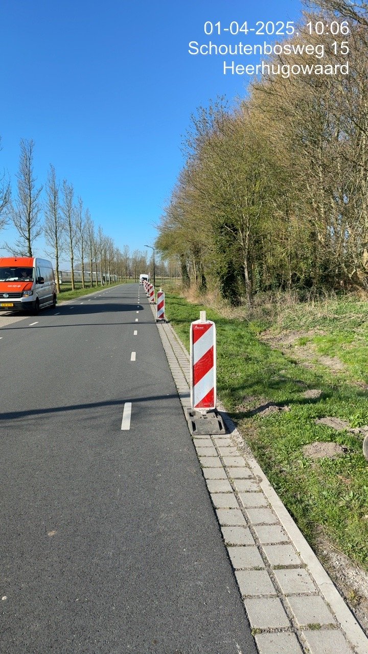 Road with construction barriers on the right side, lined with trees and a parked orange and white van, under a clear blue sky, with a timestamp and location overlay.