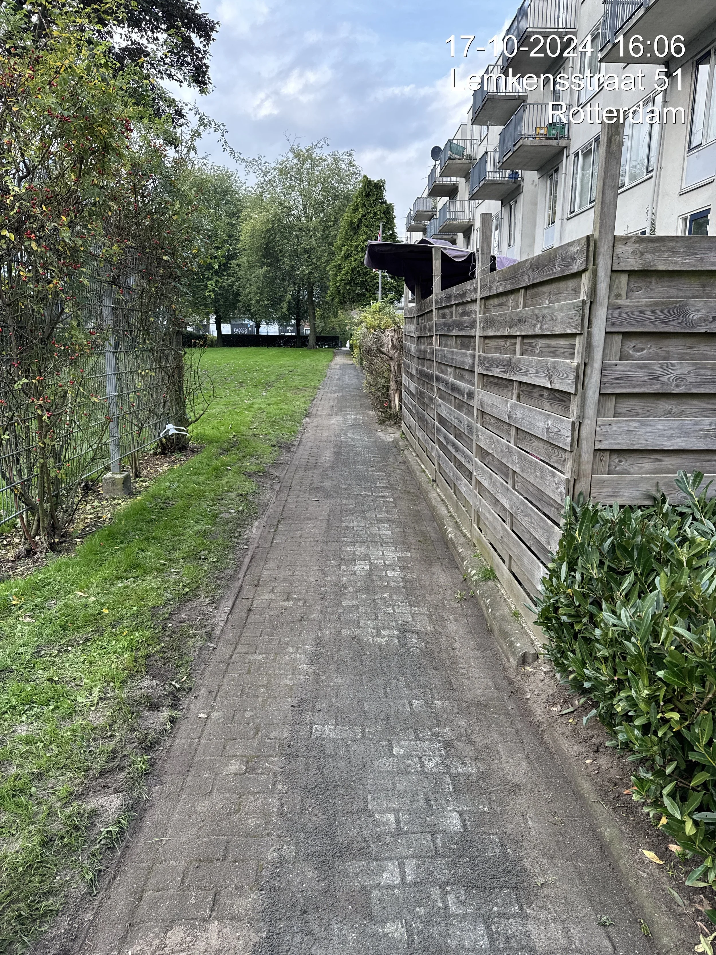 A narrow brick pathway runs between a grassy area with bushes and trees on the left and a tall wooden fence on the right. Behind the fence are apartment buildings with balconies. The sky is overcast with clouds.