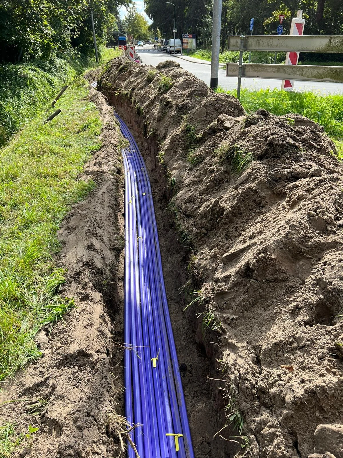 Underground utility pipes being installed in a trench alongside a road, with some grass and trees on either side.