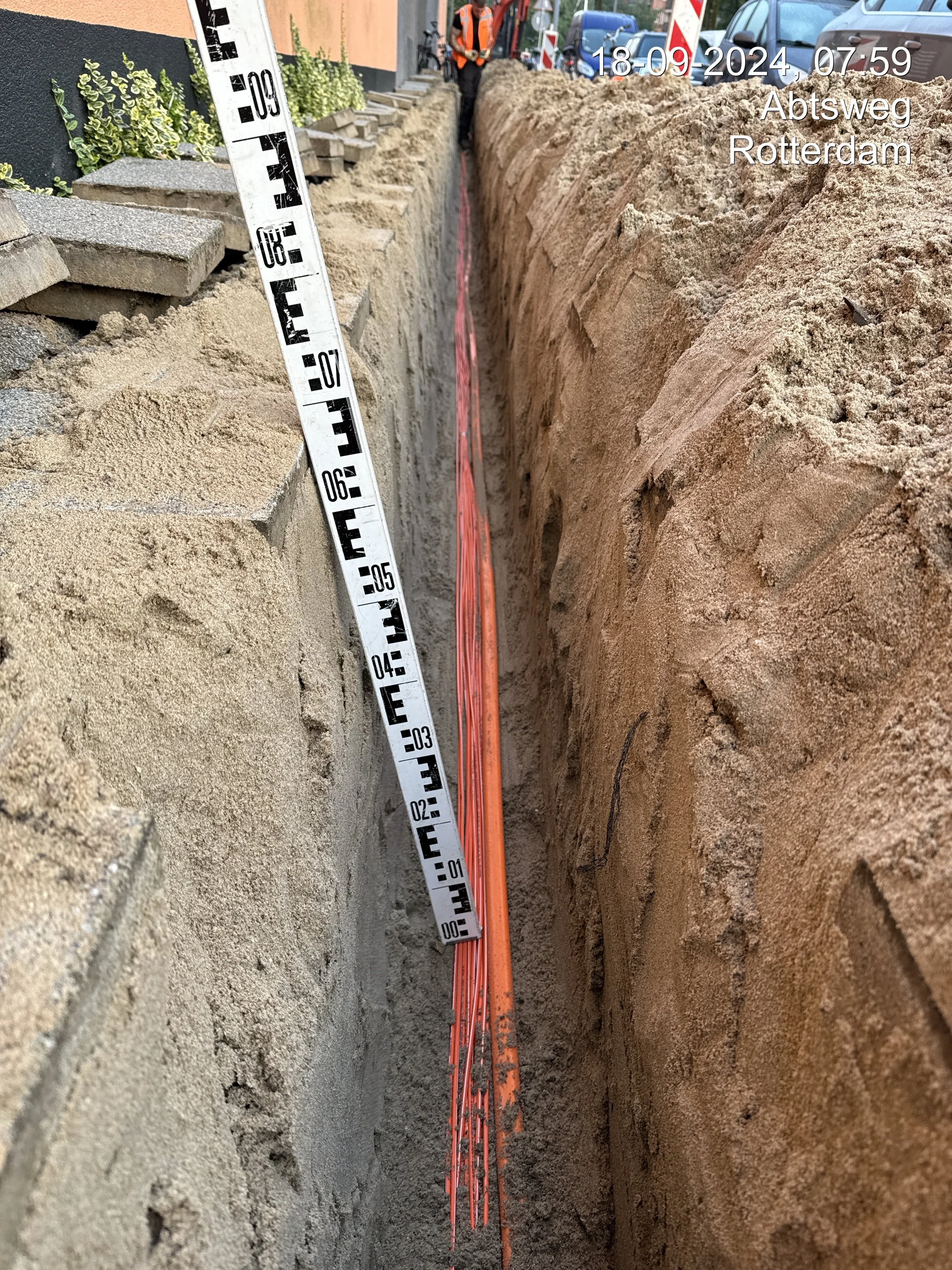 Construction site showing a trench with an orange laser level, a measuring tape, and a worker in an orange safety vest at Abtsweg in Rotterdam. Cars are parked nearby. The photo was taken on September 18, 2024, at 7:59 AM.
