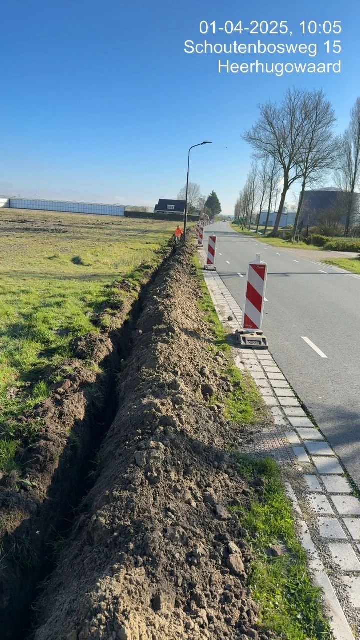 A road with a construction trench dug along the side, with barriers and a streetlamp, and trees and houses in the background.