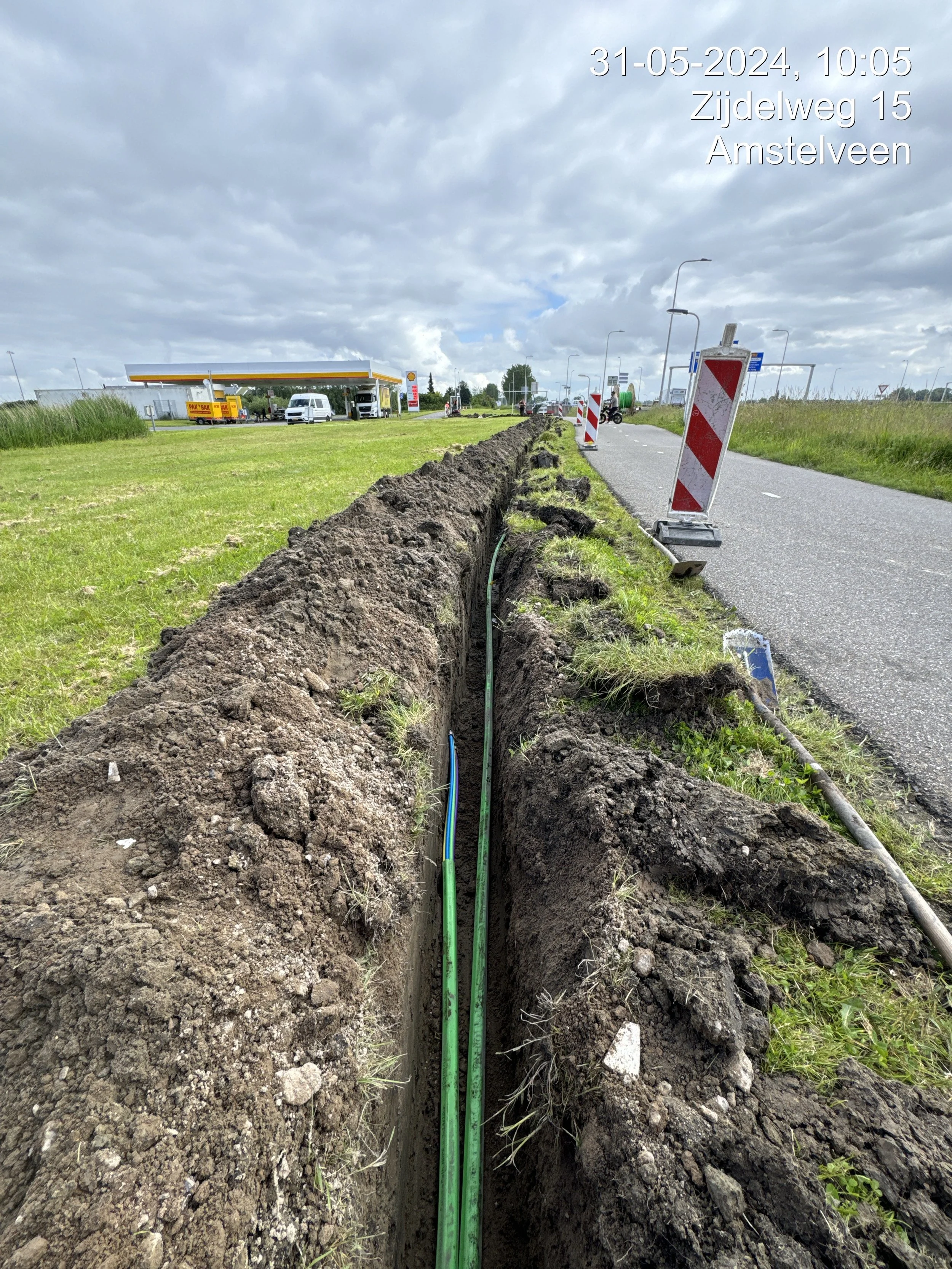 A road construction site with a deep trench dug along the side of a road, containing green and blue cables or pipes. There is a temporary barrier with red and white diagonal stripes. In the background, there is a Shell gas station and some vehicles, including a white van and motorcycles, against a cloudy sky. The image has a timestamp and location information overlaid in the top right corner.