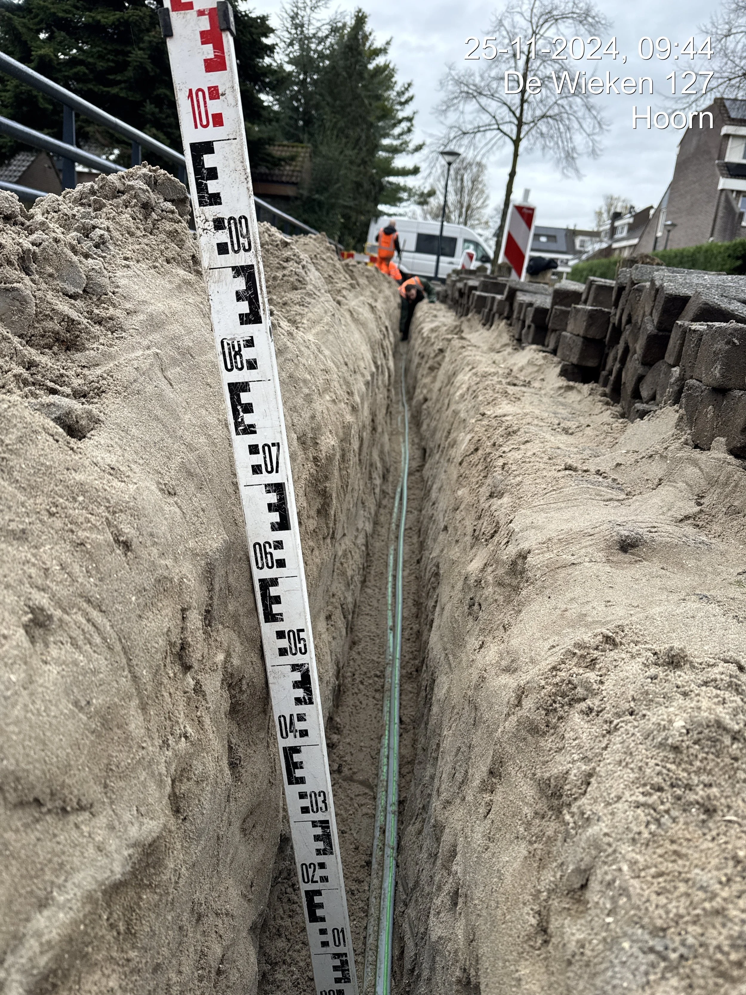 Construction workers in orange safety vests working in a trench on a residential street, with measuring equipment and a white van in the background, overcast sky.