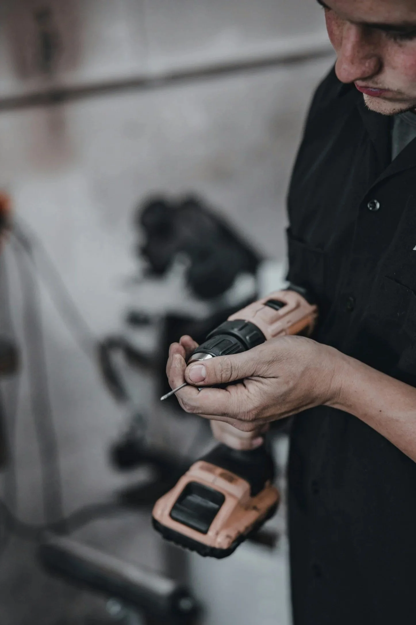 A person in black clothing holding a cordless drill in their hands, with a phone lying on a metal surface nearby.