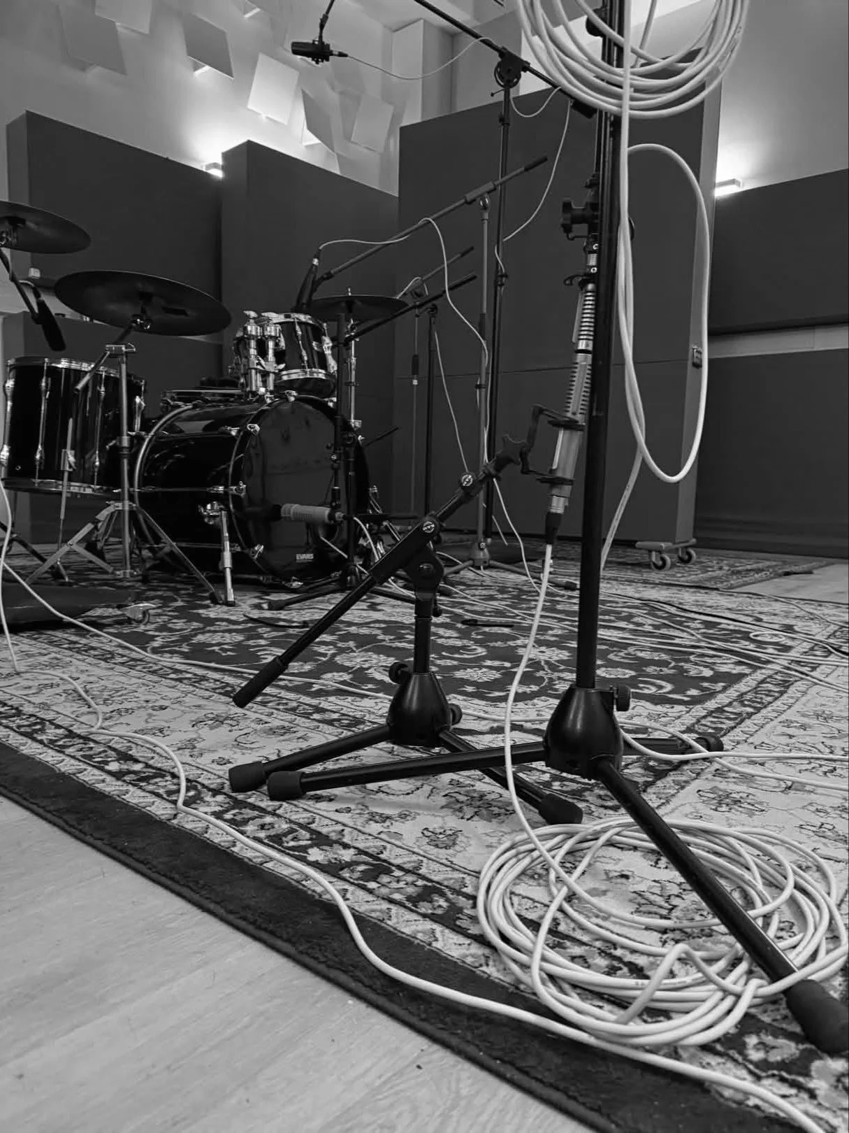 In a recording studio, a drum set is positioned on a patterned rug, surrounded by multiple microphones on stands and connected with several cables. The background features acoustic panels and studio equipment.
