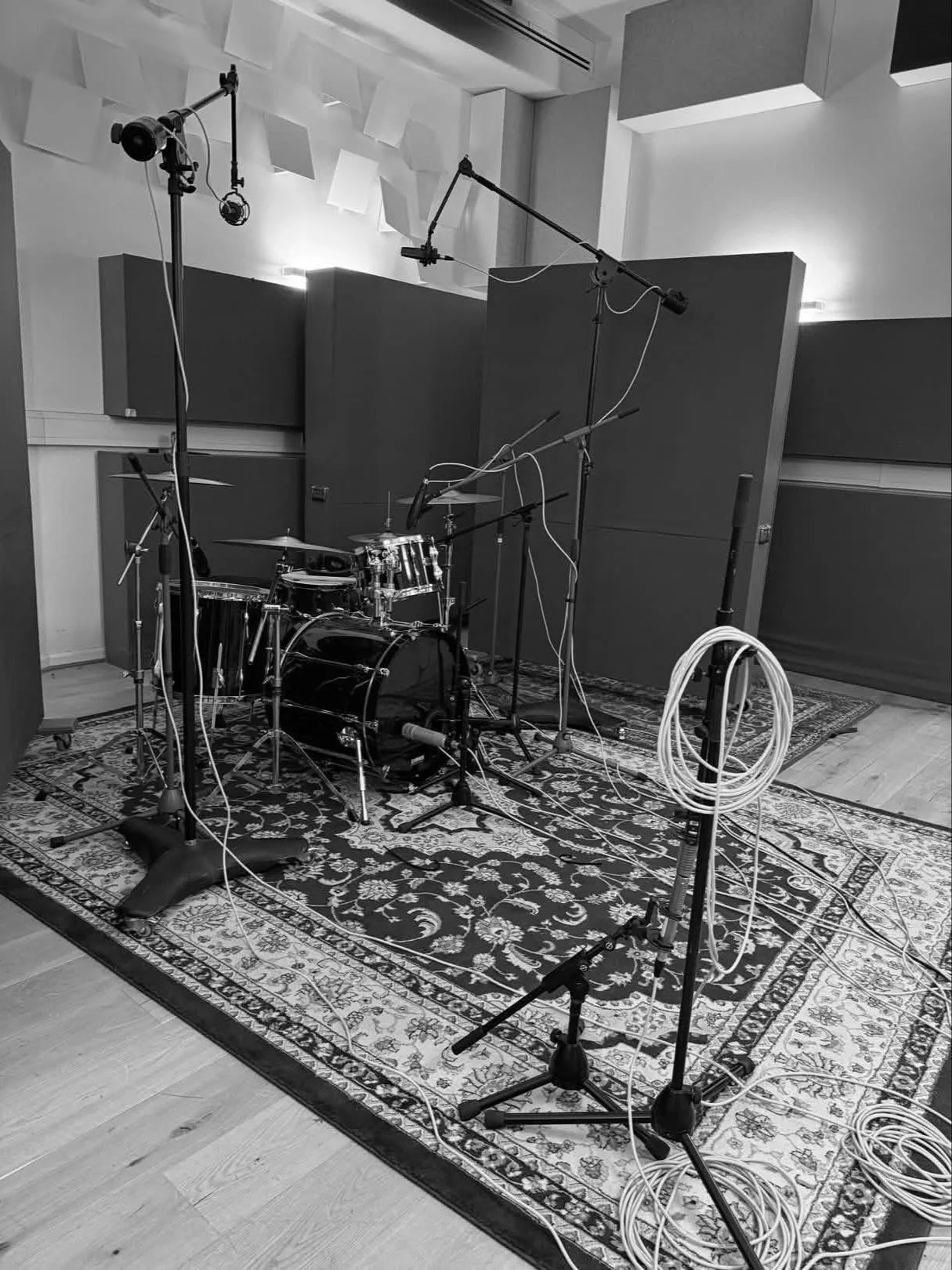 A black and white photo of a music recording studio setup with drums, multiple microphones, and cables on a patterned rug.
