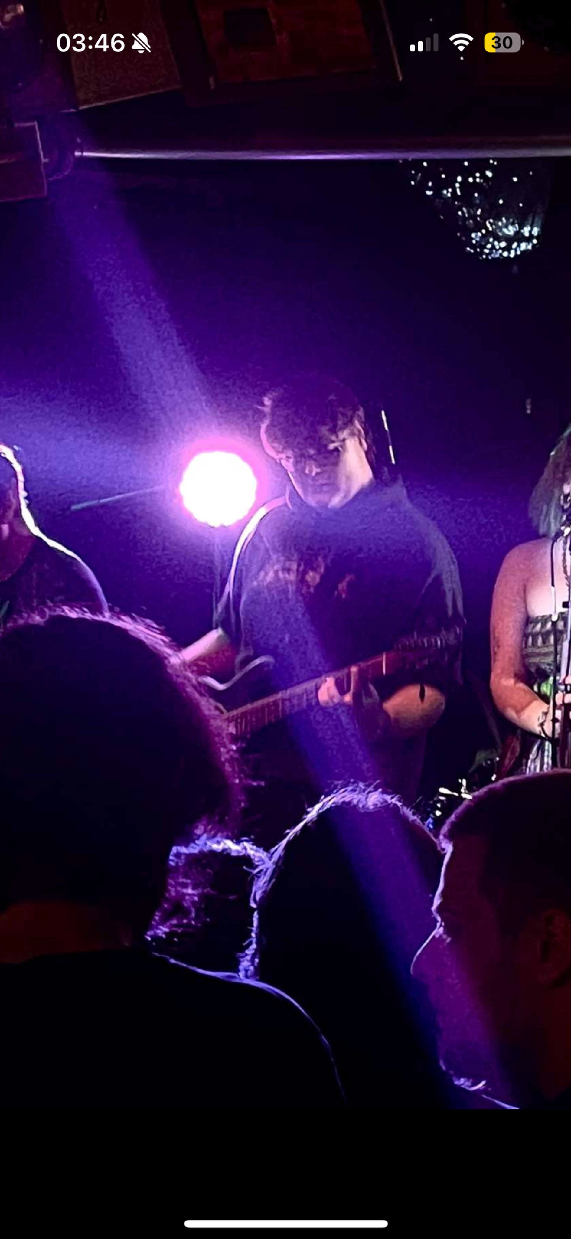 A person playing guitar on stage under purple and pink lighting, with audience members visible in the foreground and a disco ball hanging from the ceiling.