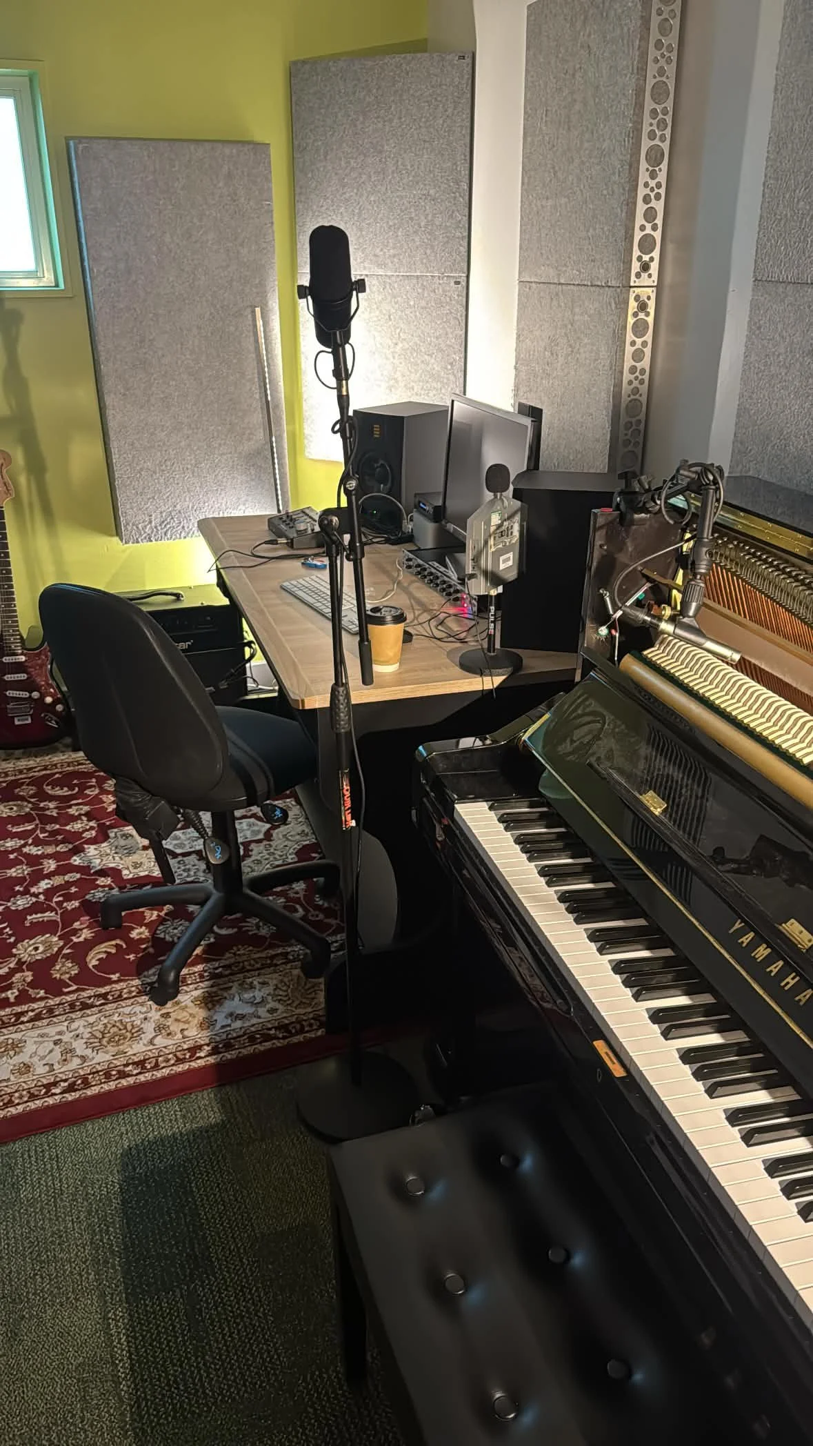 A music recording studio with a piano, a computer, studio monitors, a microphone, a chair, acoustic panels on the walls, and a red patterned rug.