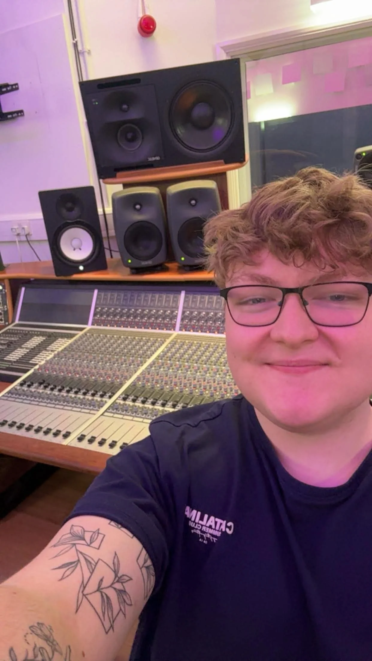 A young man with glasses and tattoos on his arm taking a selfie in a recording studio. Behind him is a large mixing console and multiple studio monitor speakers, with a large speaker and additional monitors on stands against the wall.
