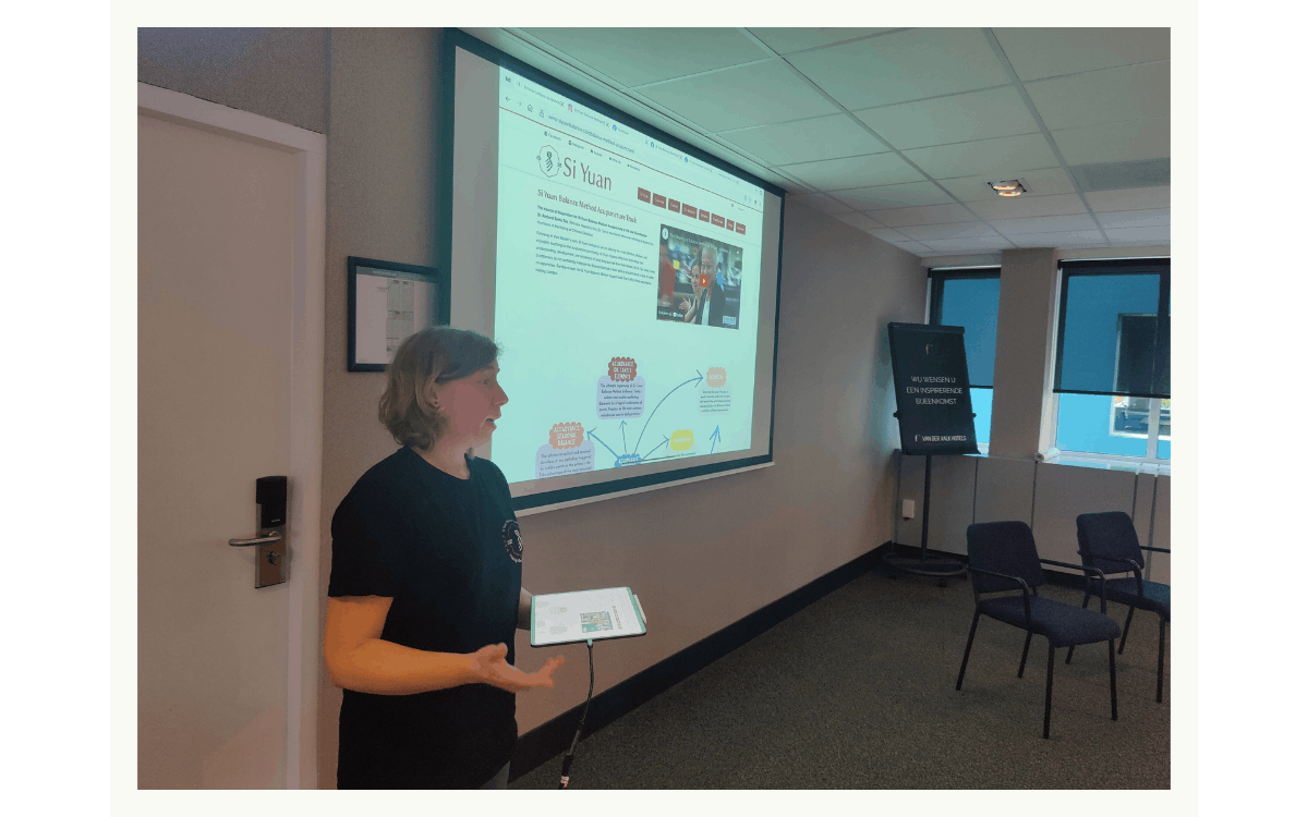Woman presenting in a conference room in front of a projection screen displaying a webpage, holding a tablet, with chairs and windows in the background.
