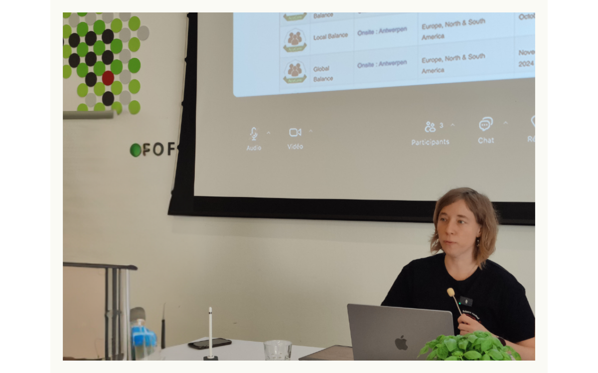 A woman sitting at a table with a laptop, microphone, and a small green plant in front of her, giving a presentation in a conference room. There is a large screen behind her displaying a presentation slide with text and icons.