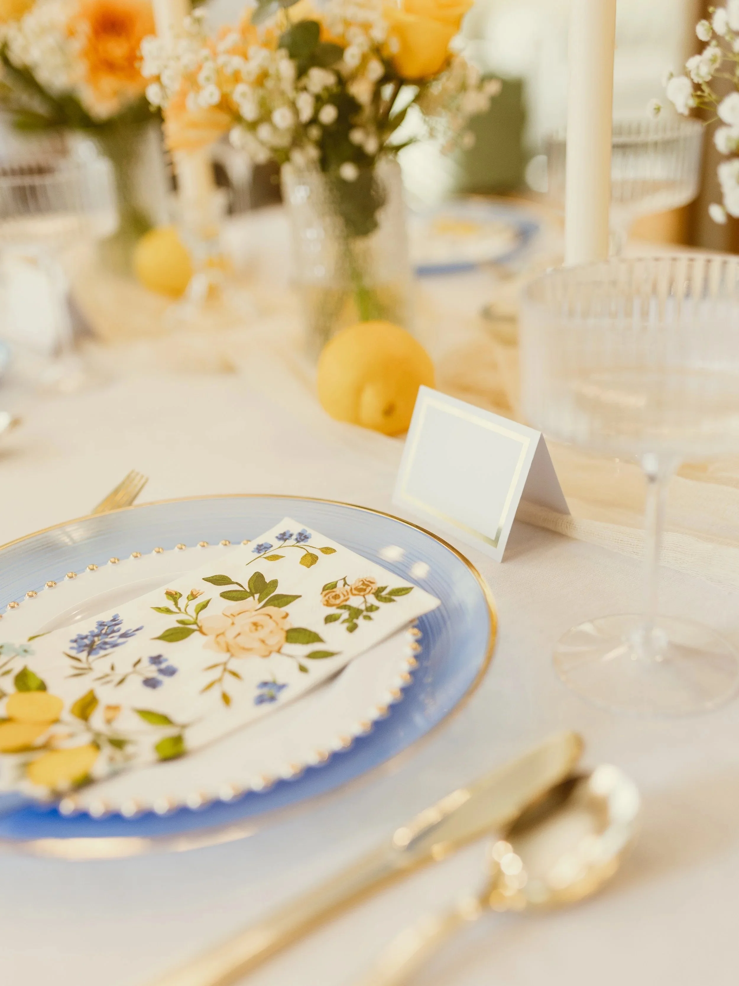 Elegant table setting with floral-patterned plate, floral centerpiece, candles, and a gold-rimmed napkin.