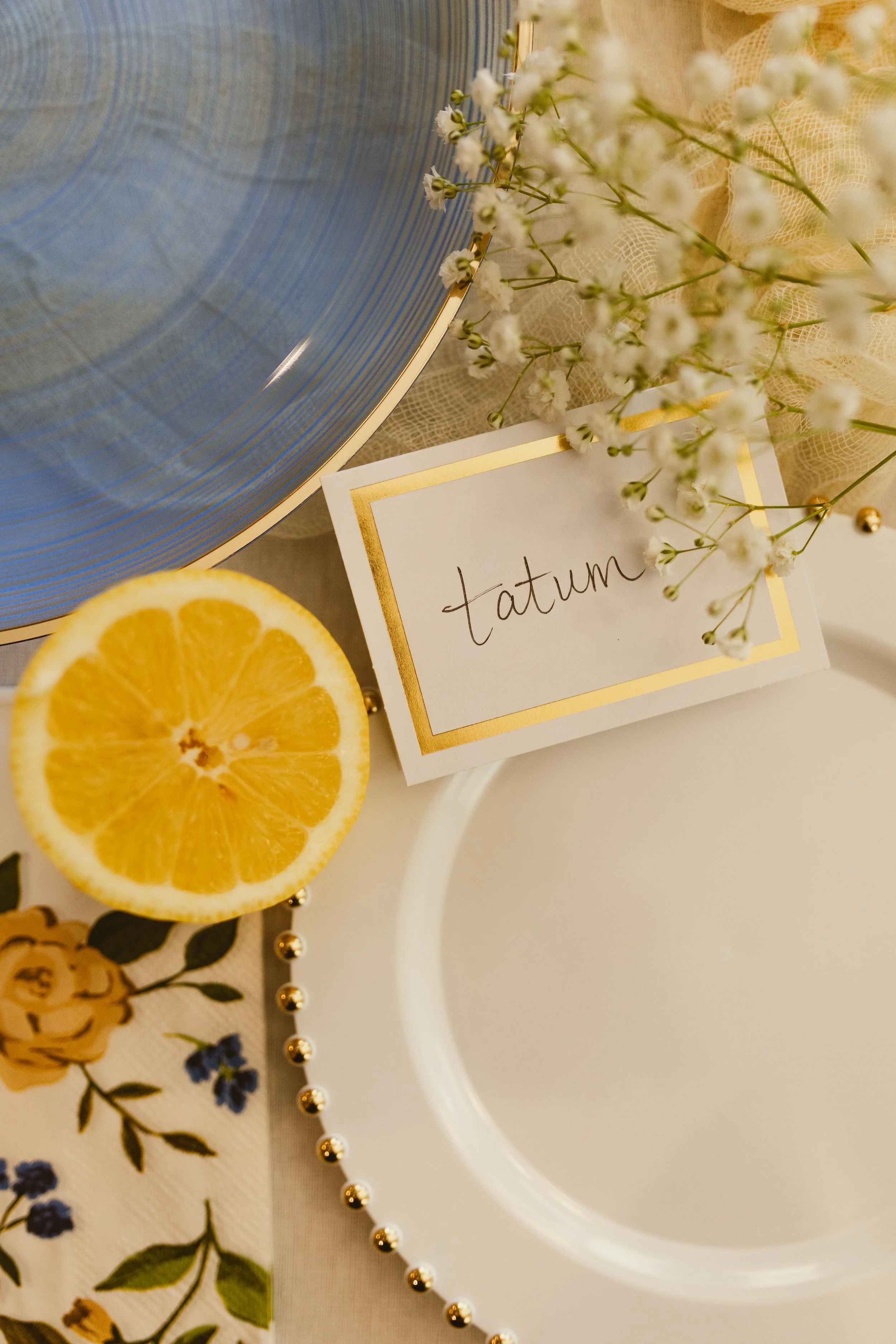 A close-up image of a lemon slice, a white card with the word 'tatum' written on it, white baby's breath flowers, a blue decorative plate, a string of gold beads, a floral napkin, and part of a white plate.