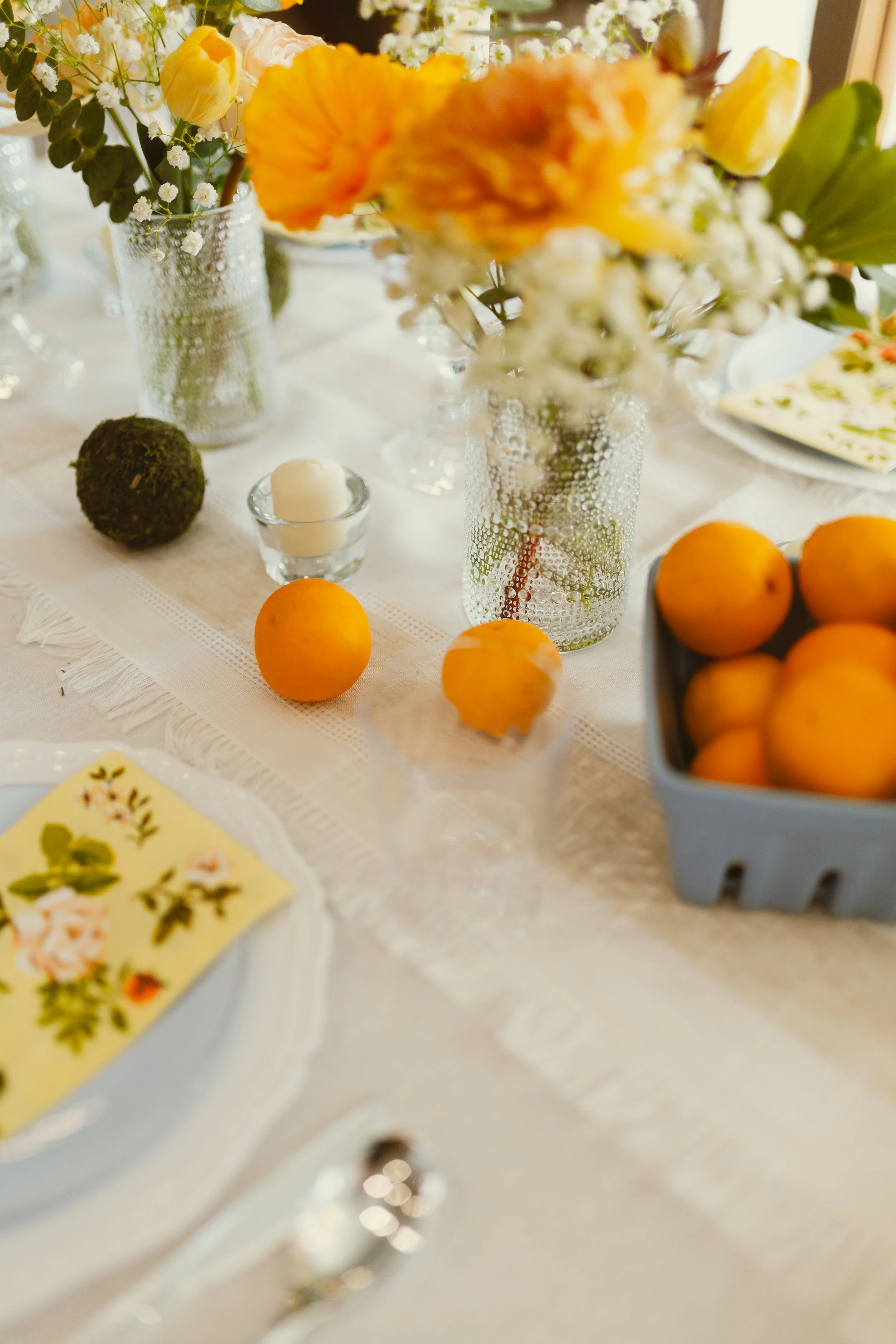 Close-up of a table with floral arrangements in glass vases, a small candle in a glass holder, and a few oranges. The background includes patterned plates and more flowers, with a soft, warm light illuminating the scene.