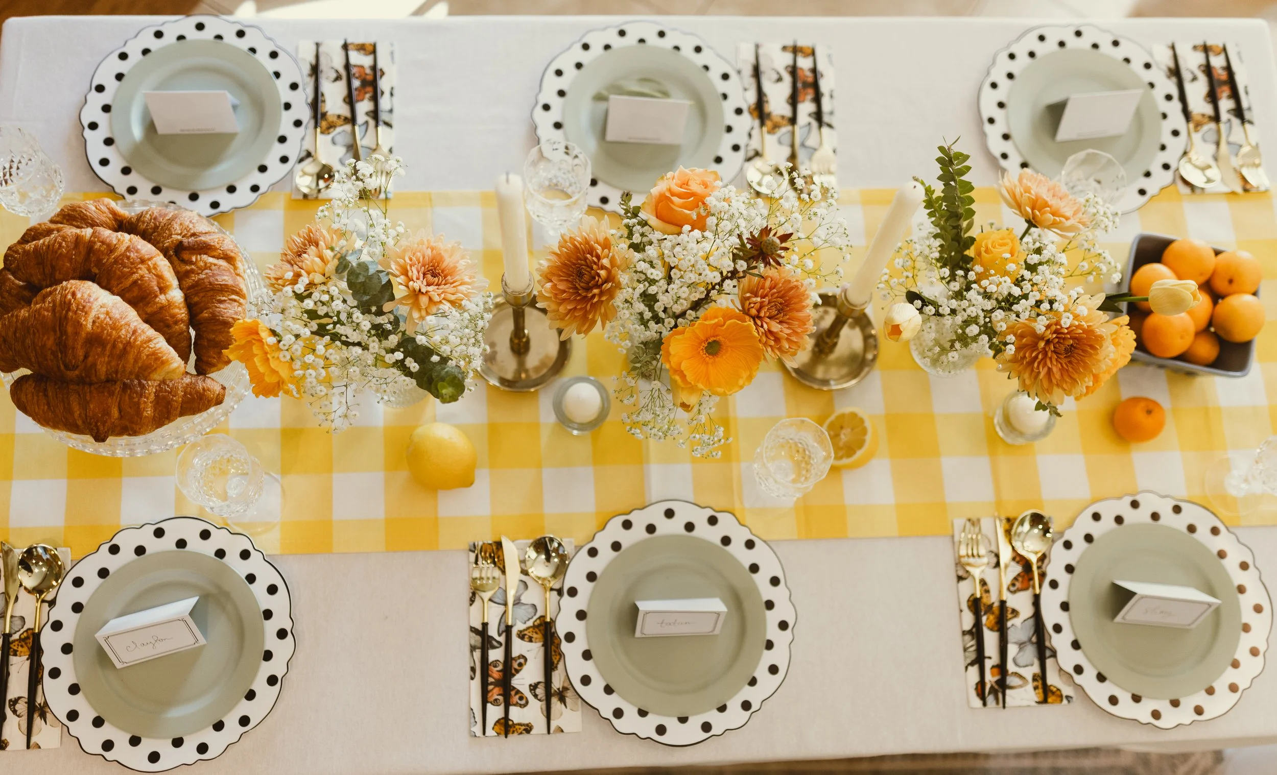 A table setting with floral centerpiece, croissants on a glass cake stand, lemons and oranges, polka dot plates, gold utensils, and a yellow checkered table runner.