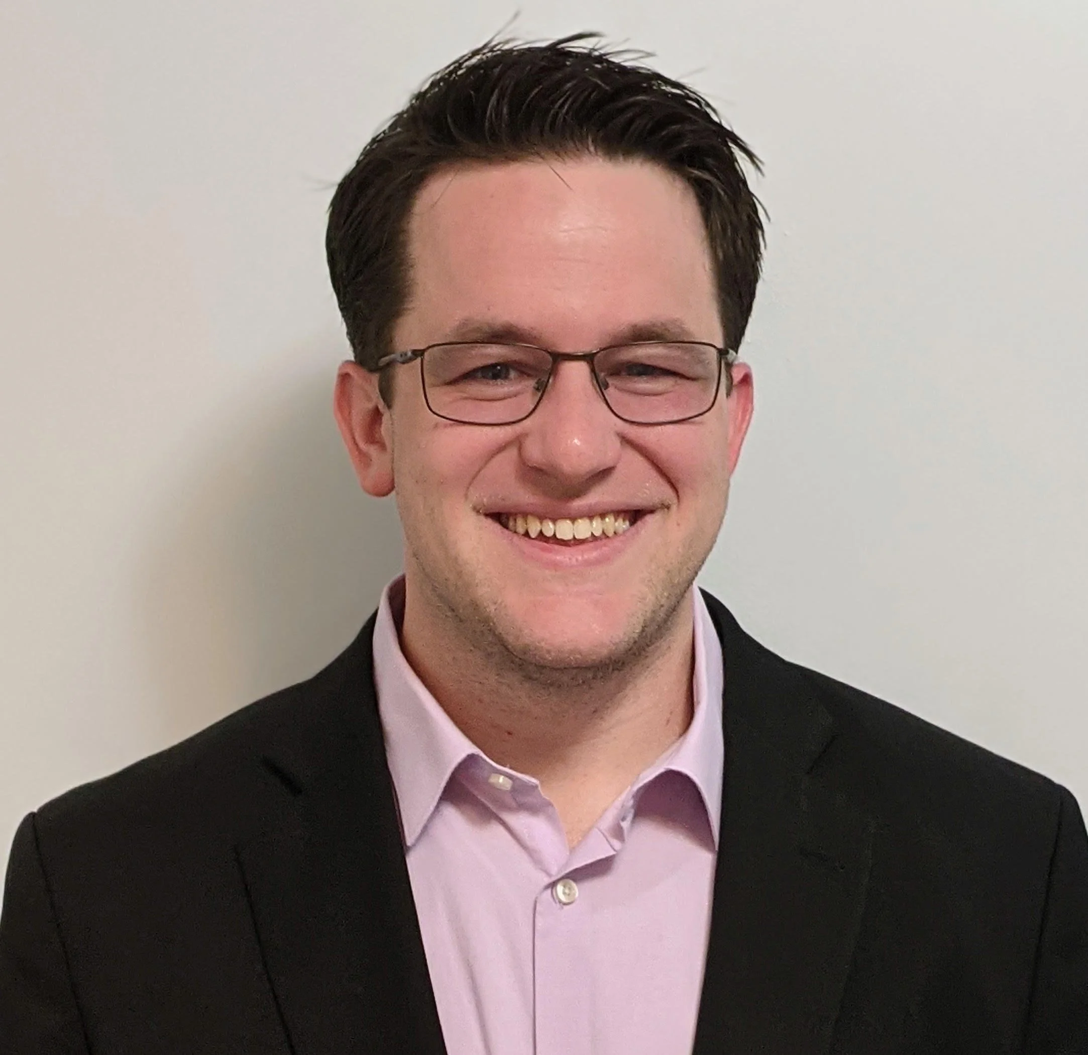 Close-up photo of a young man with dark hair, glasses, wearing a pink shirt and black blazer, smiling against a plain white background.
