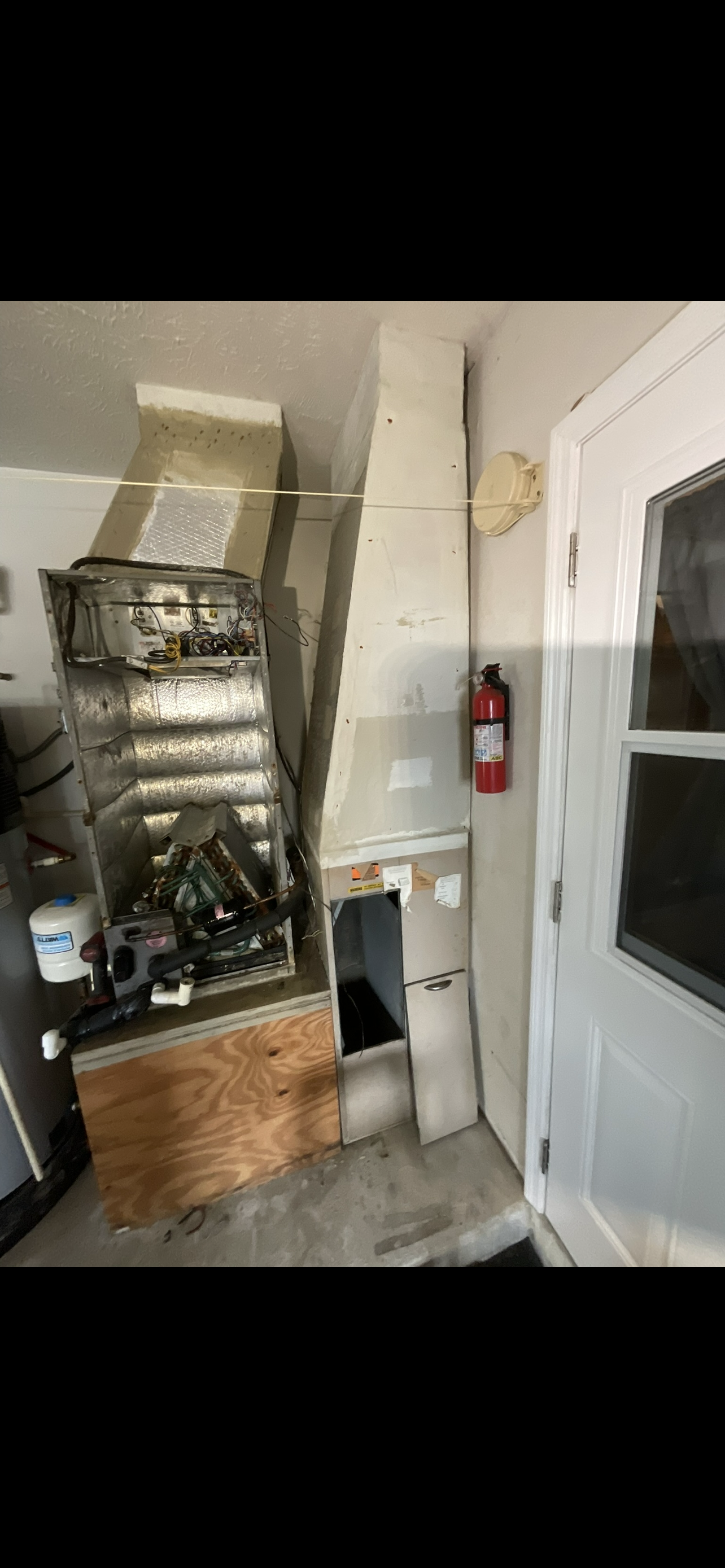 Disassembled furnace with exposed components, a fire extinguisher mounted on the wall, and a white door with a window.