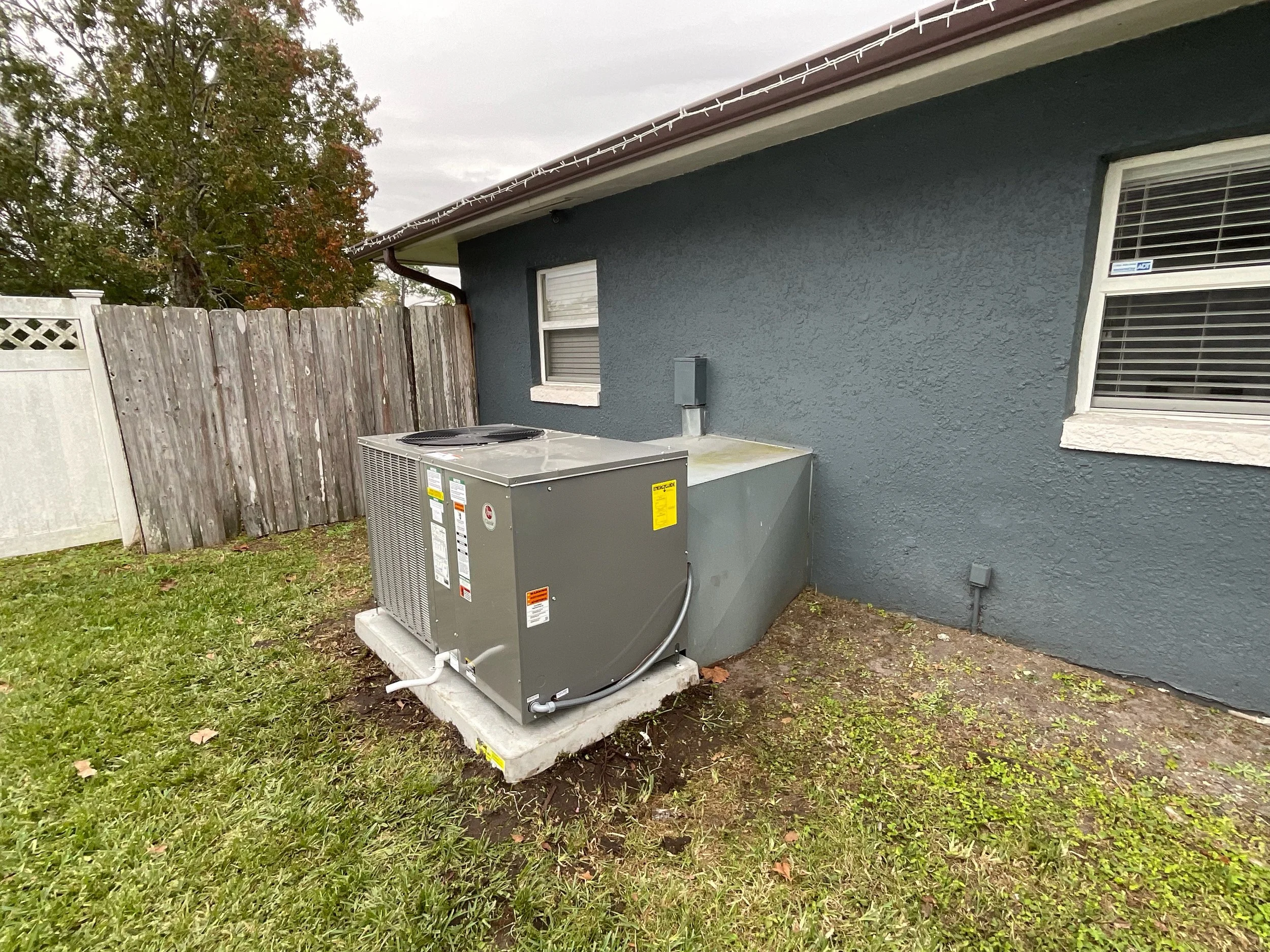 The image shows the side of a house with gray textured walls, two windows with closed blinds, a gray utility box attached to the wall, and an air conditioning unit on a concrete pad in the yard. A weathered wooden fence runs along the left side, and a large tree with autumn-colored leaves is visible in the background. There are Christmas lights hanging along the roof edge and some grass in the yard.