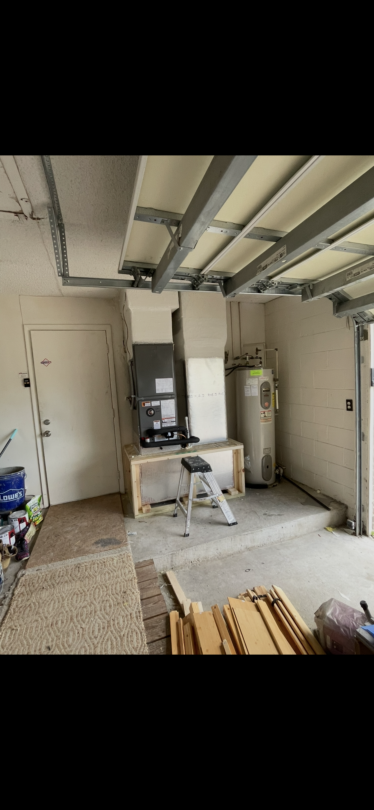 A garage with an unfinished garage door, a step ladder, water heater, workbench, and various construction materials.