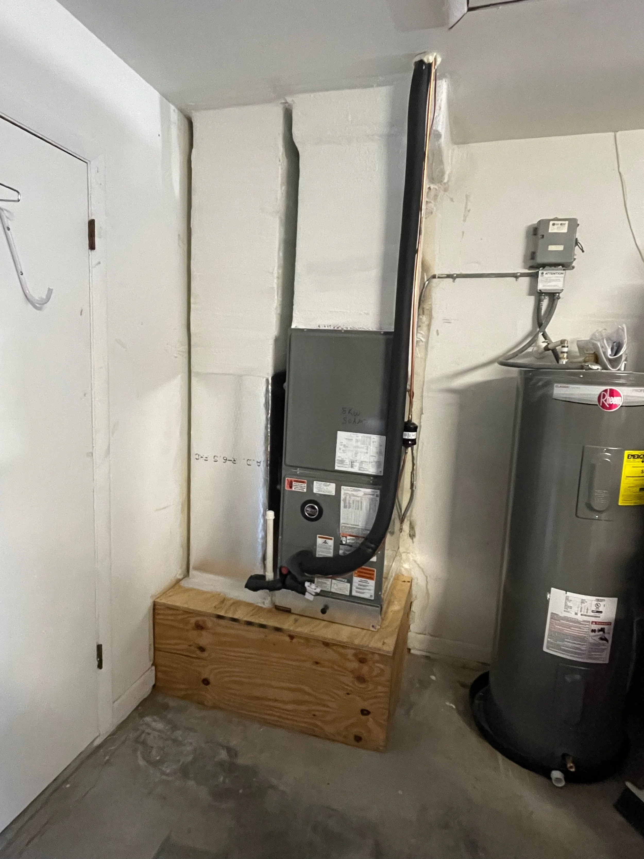 A basement utility room with a white door on the left, a white painted wall, and a concrete floor. In the center, a furnace is placed on a wooden platform with a black duct connected to it. To the right, there is a water heater with a gray exterior and yellow energy sticker, with pipes and wires attached.