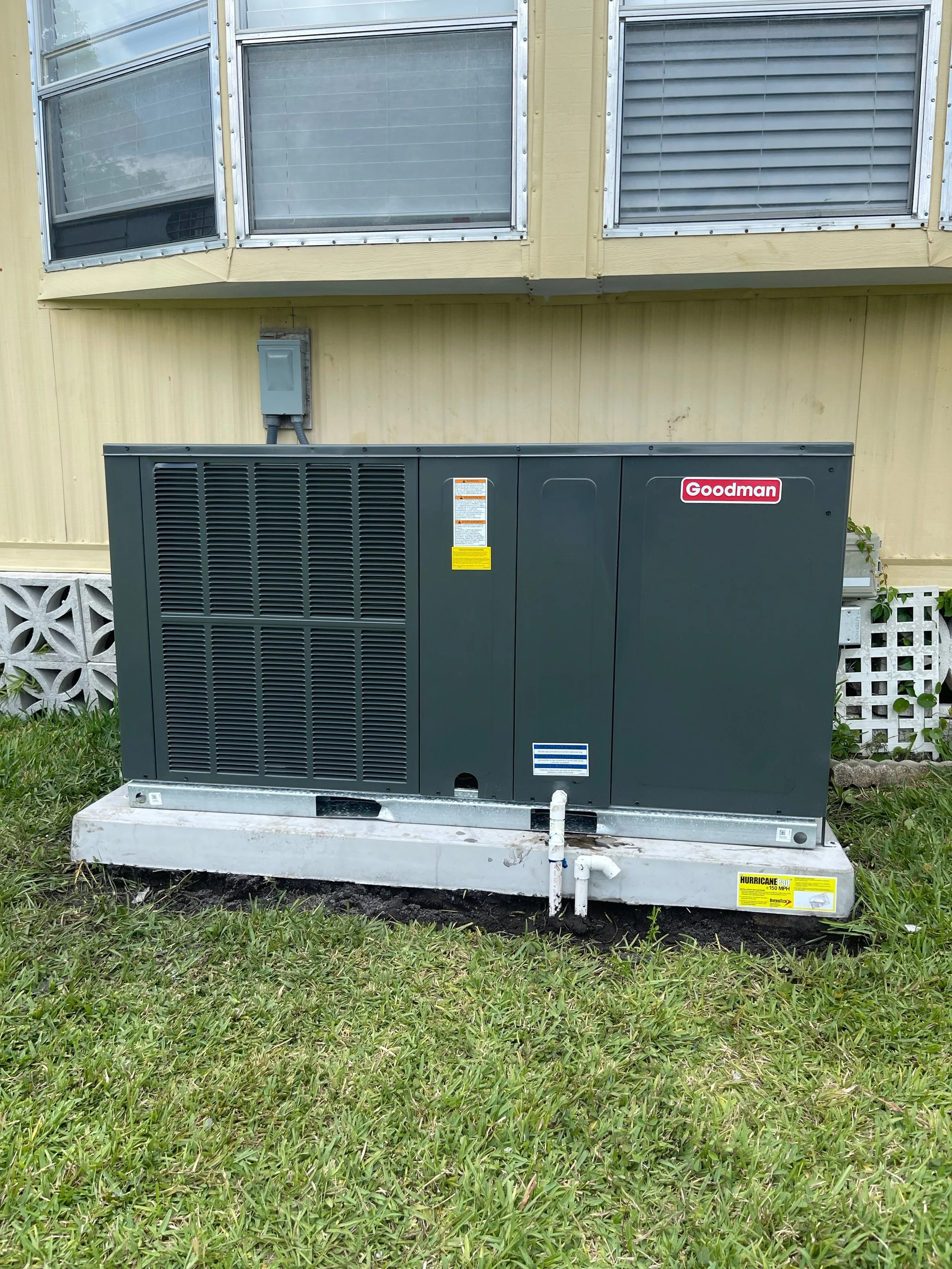 A dark gray Goodman air conditioning unit installed on a concrete slab outside a yellow house with a second story. The house has windows with blinds, some open. There are white lattice and concrete blocks behind the unit, and pipes are connected to the unit. The grass in front is green.