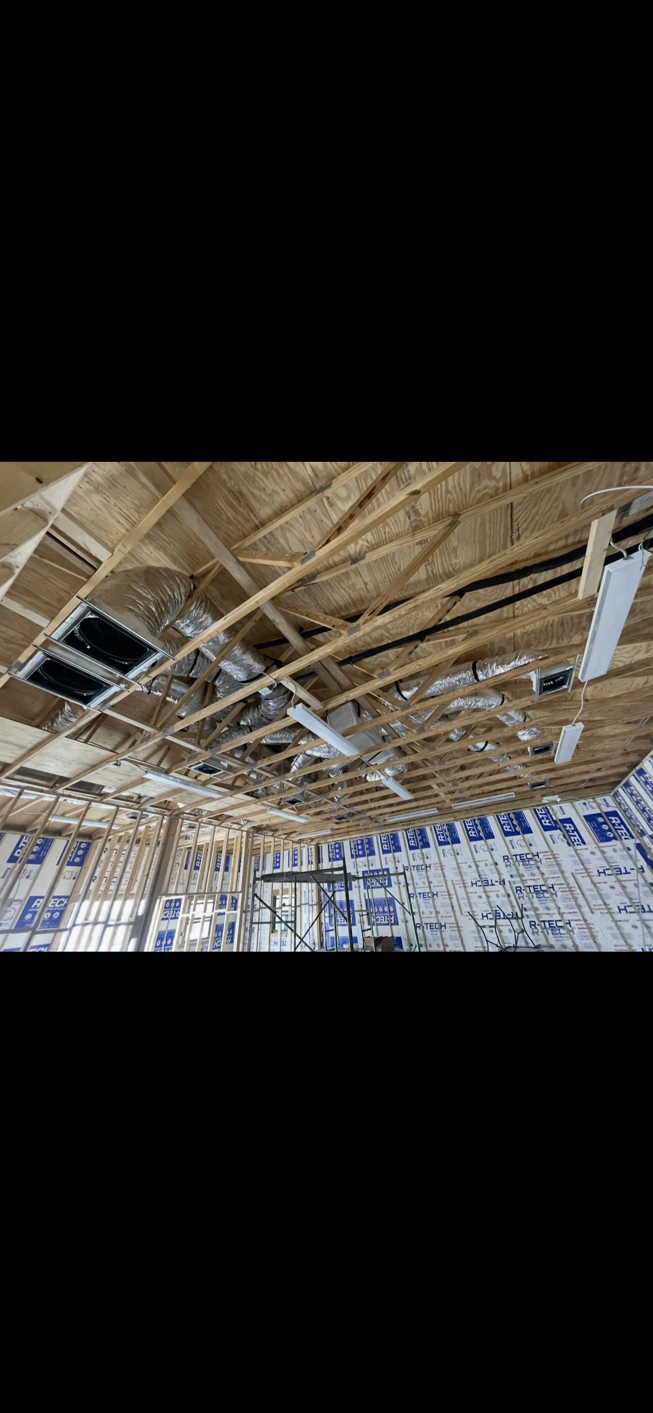 Construction site with exposed wooden ceiling framing and ductwork, insulation, and lighting fixtures visible inside a building under construction.