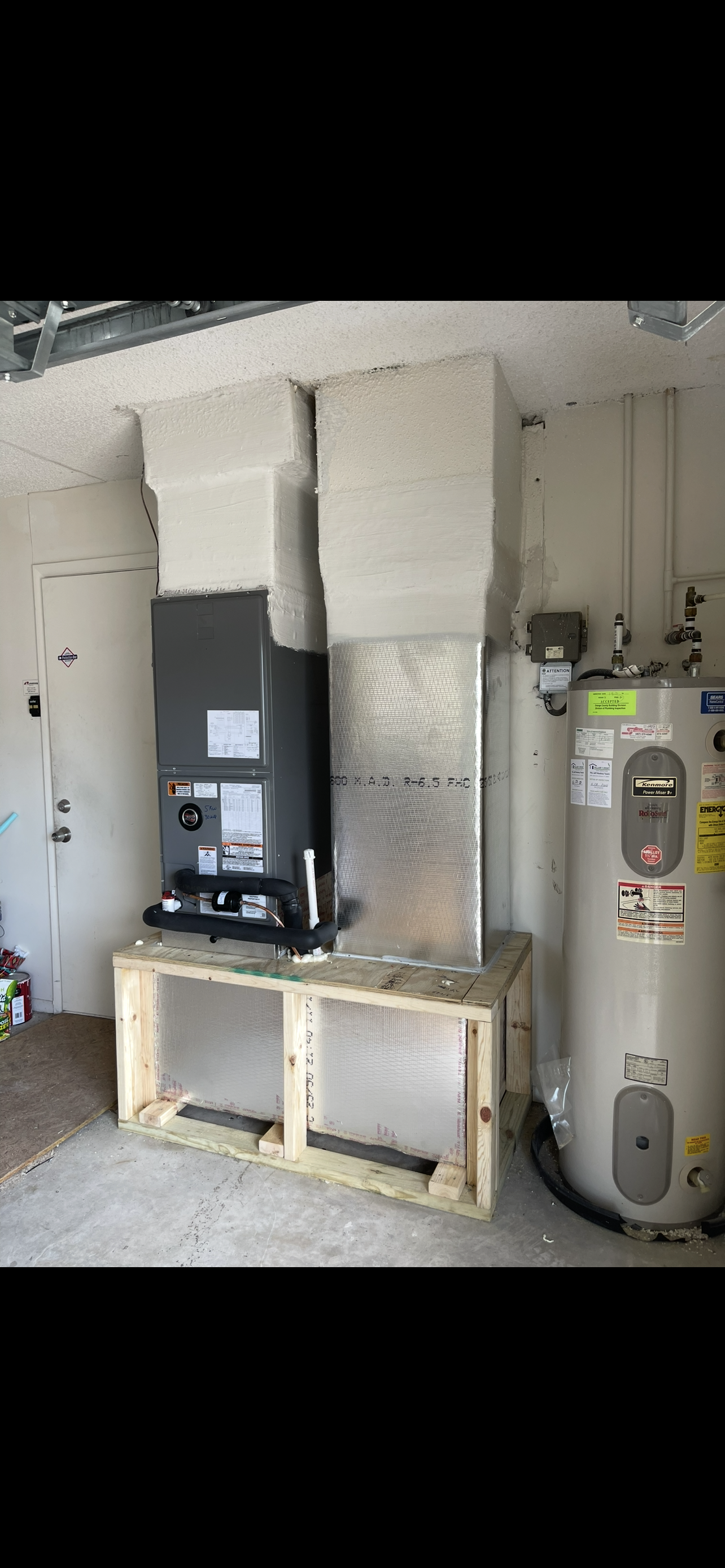 A utility room with a large water heater on the right, an HVAC unit on the left, and insulation work in progress on a wooden frame in the center.