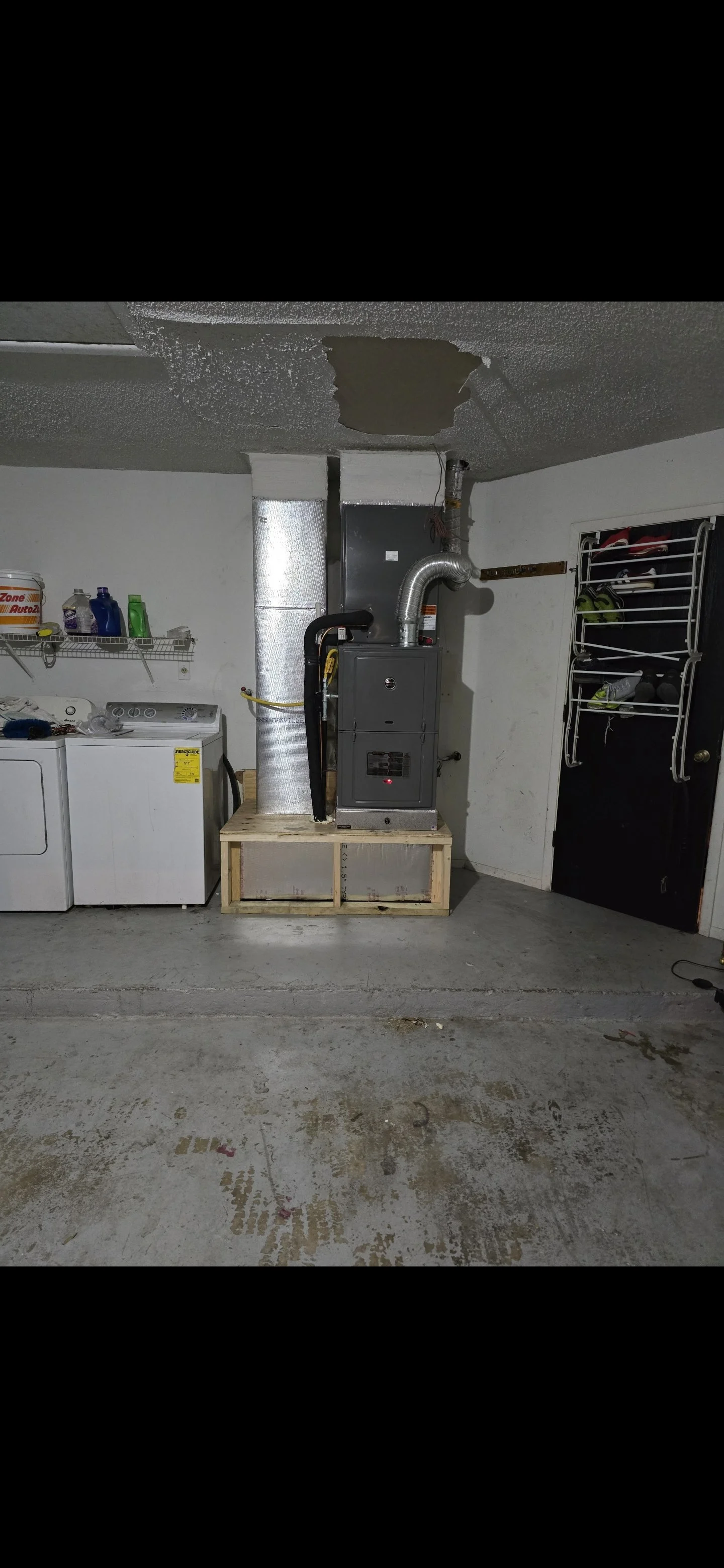 Garage with a washing machine, laundry supplies, and a furnace on a wooden platform, with a piped vent and a door with shoe storage racks.