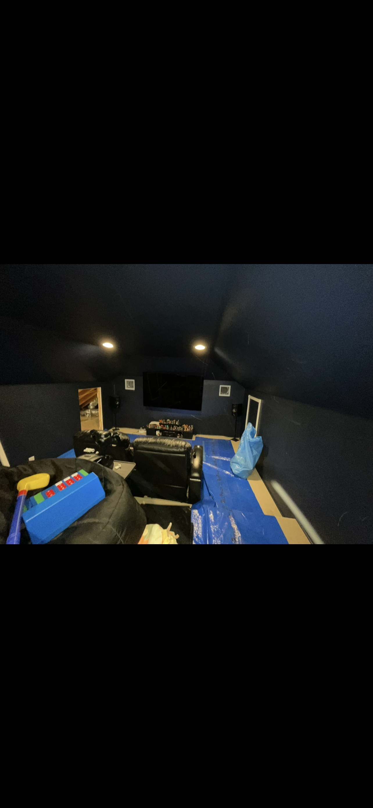 A home theater room with a large flat screen TV, black leather armchairs, and a blue tarp on the floor. There are gaming controllers and a blue toy on a black sack in the foreground, and a blue plastic bag on the right side of the room.