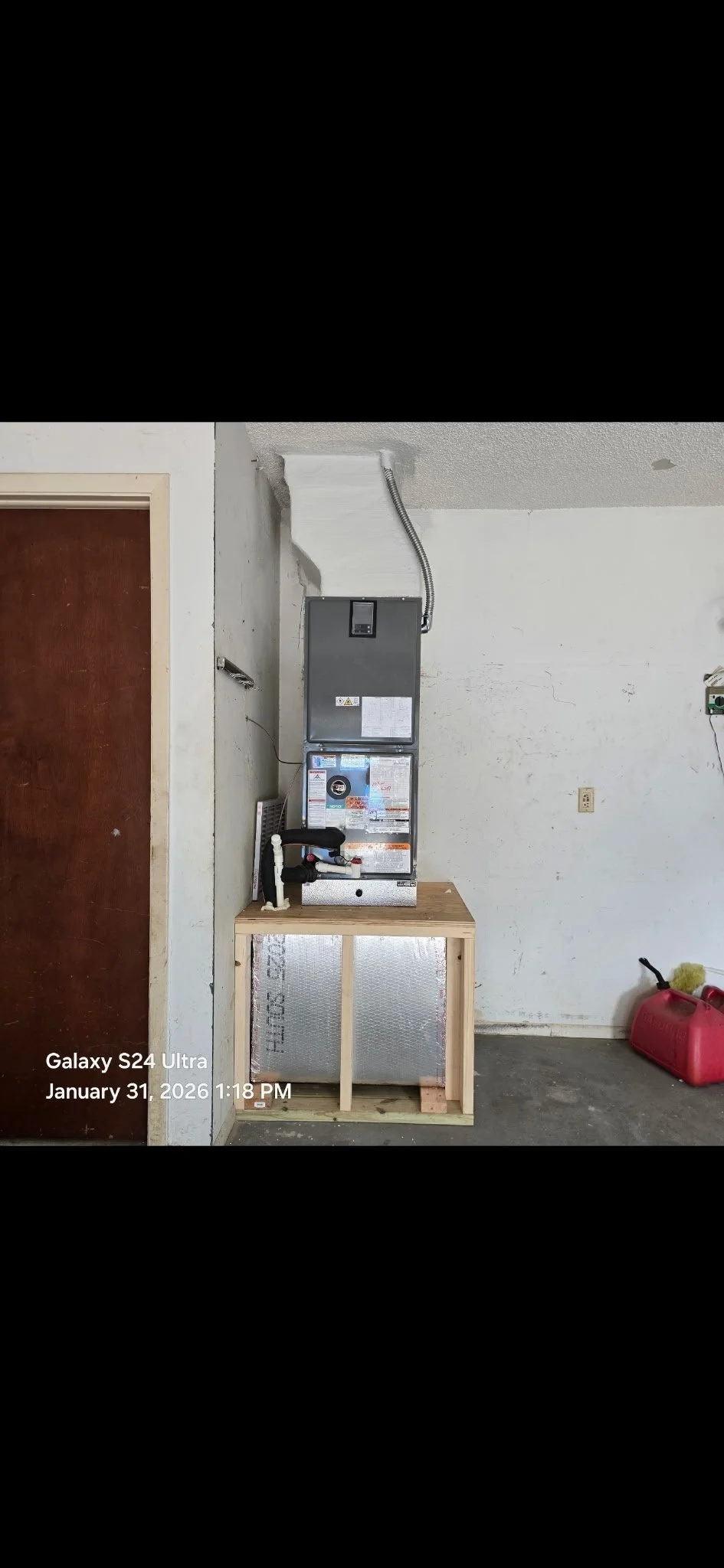 An HVAC unit installed on a wooden stand inside a garage. The garage has a concrete floor, a white wall with some cracks, and a brown door on the left. There is also a red fuel container with a hose and brush on the right side.