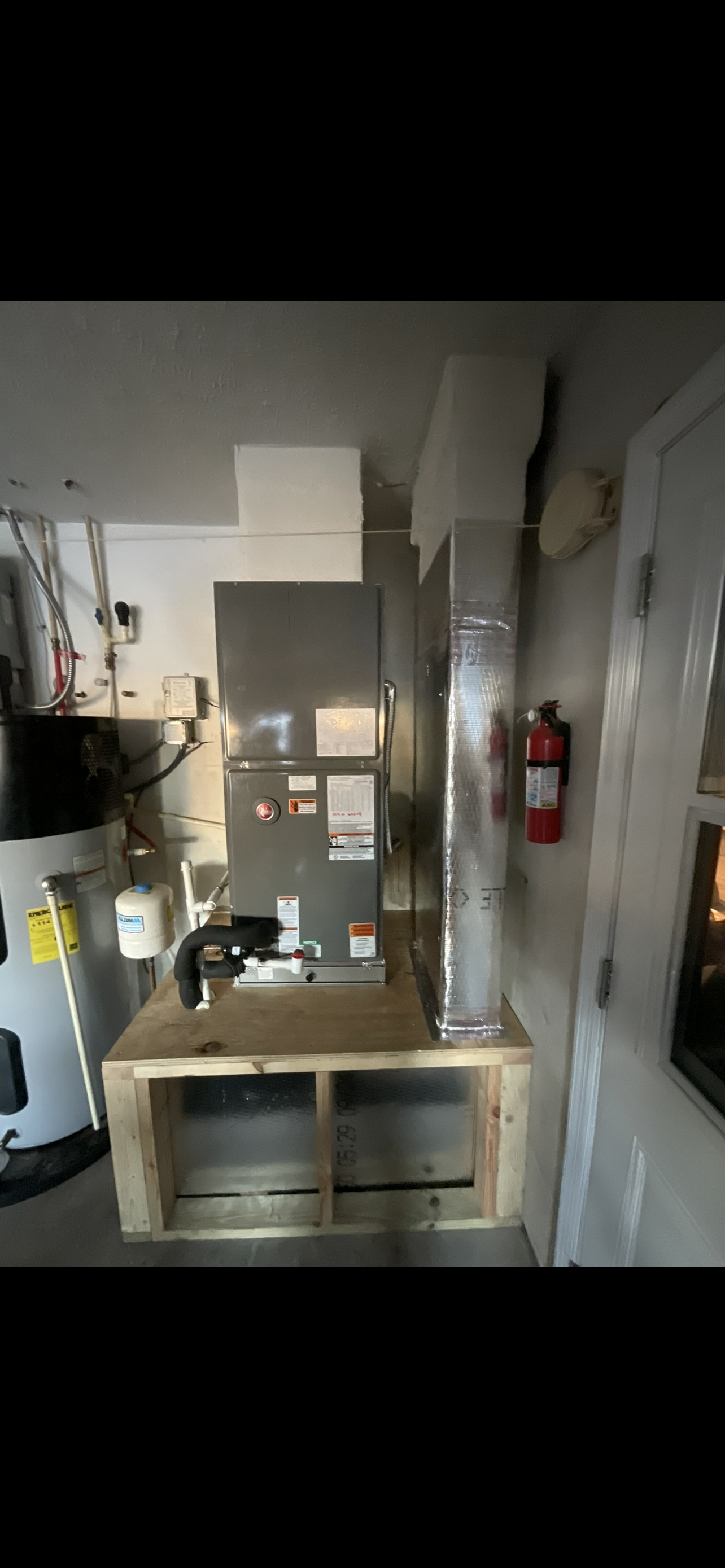 The image shows a utility or mechanical room with an HVAC unit on a wooden platform, a water heater, and a fire extinguisher mounted on the wall.
