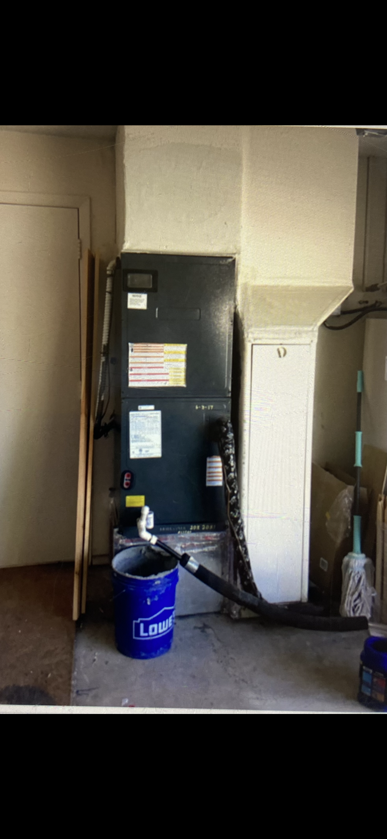 An HVAC furnace unit installed in a utility closet with a blue Lowe's bucket and cleaning tools nearby.