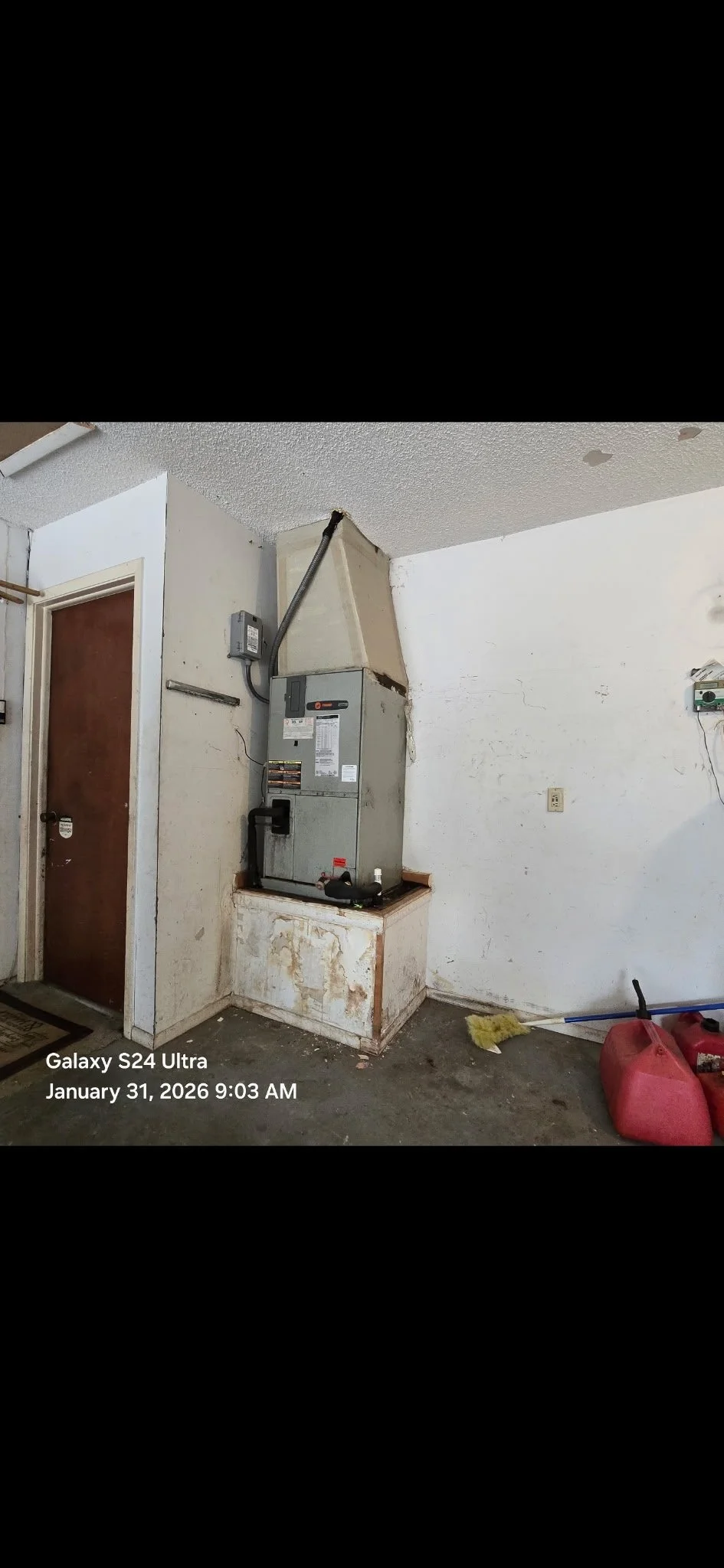 A worn, dirty corner of a room with an old heating or furnace unit installed on a small platform against the wall, with exposed wiring. There's a doorway with a closed brown door, and a white wall with some damage and cracks. On the floor, there is a broom, a red fuel container, and a pink fuel container.