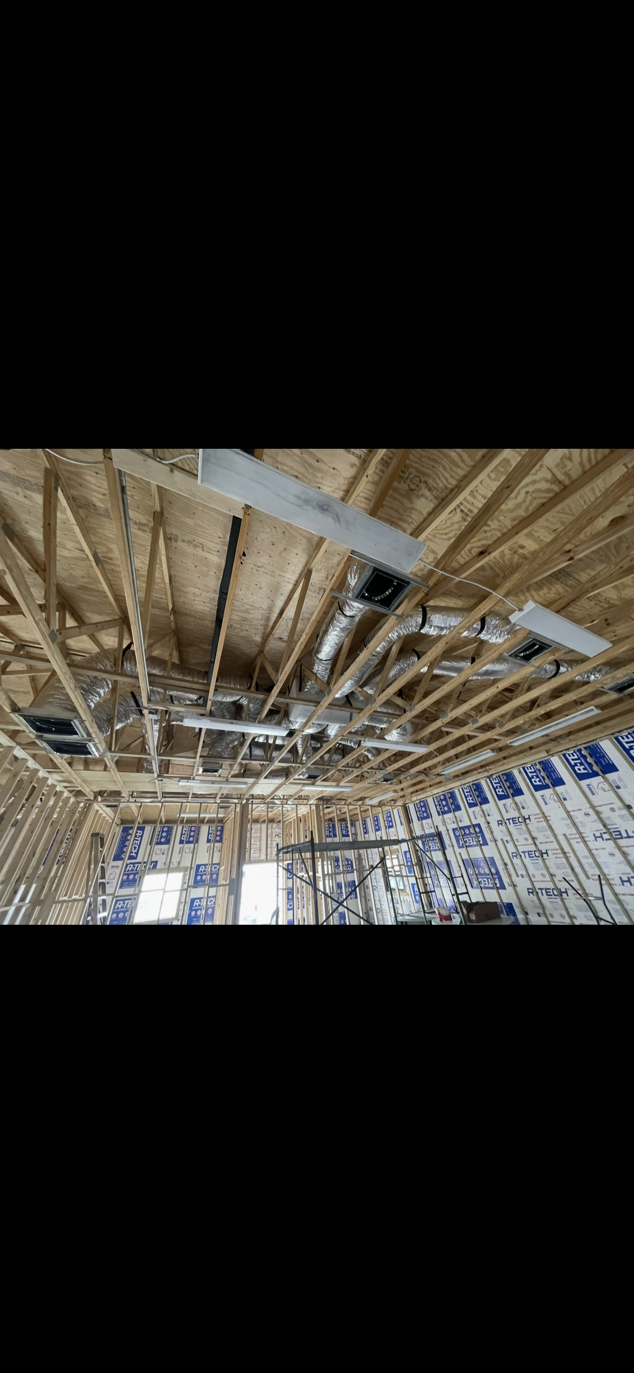 Construction site with exposed wooden framing, ductwork, and lighting fixtures on the ceiling. Insulation material on the walls, with windows allowing natural light.