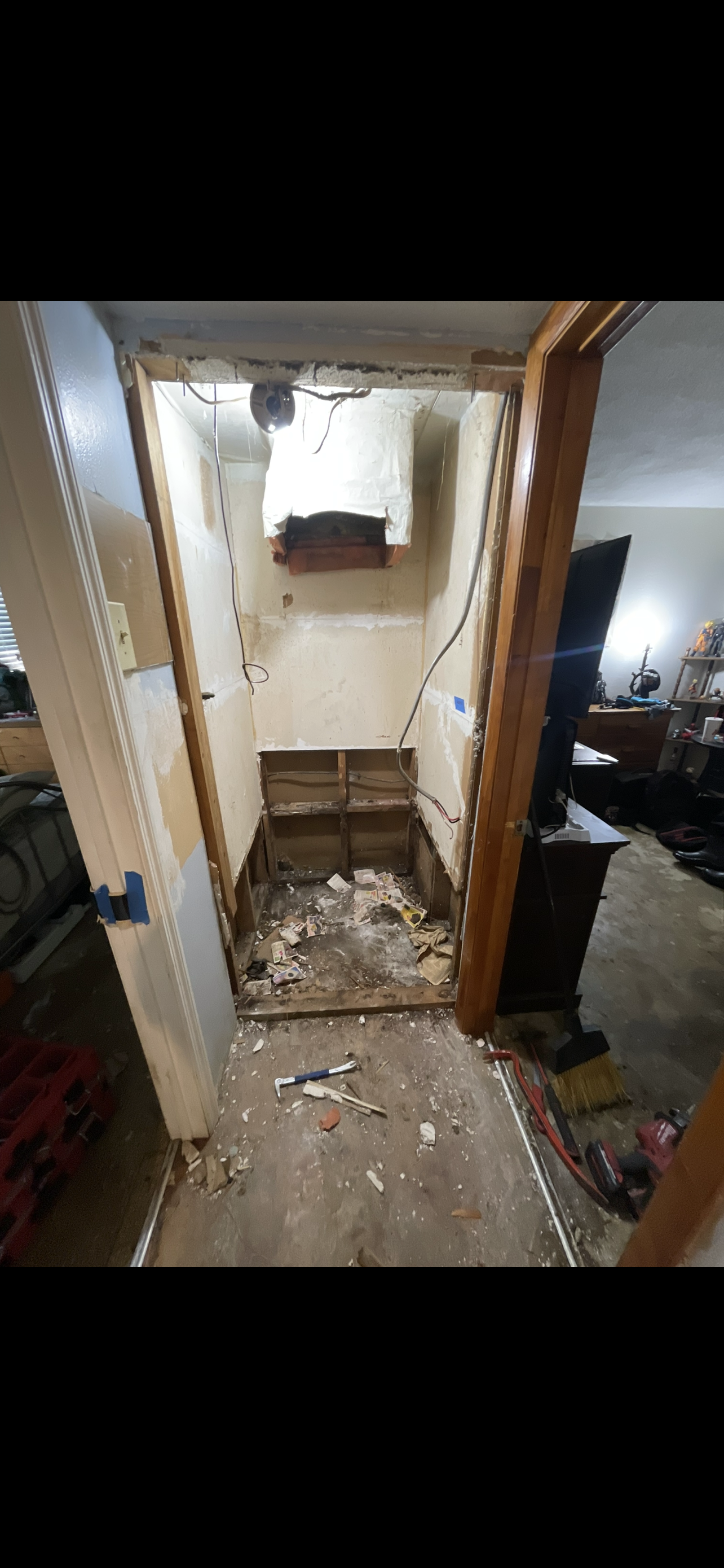 A partially demolished closet with exposed framing, debris on the floor, and a vent near the ceiling. Tool and construction materials scattered around.