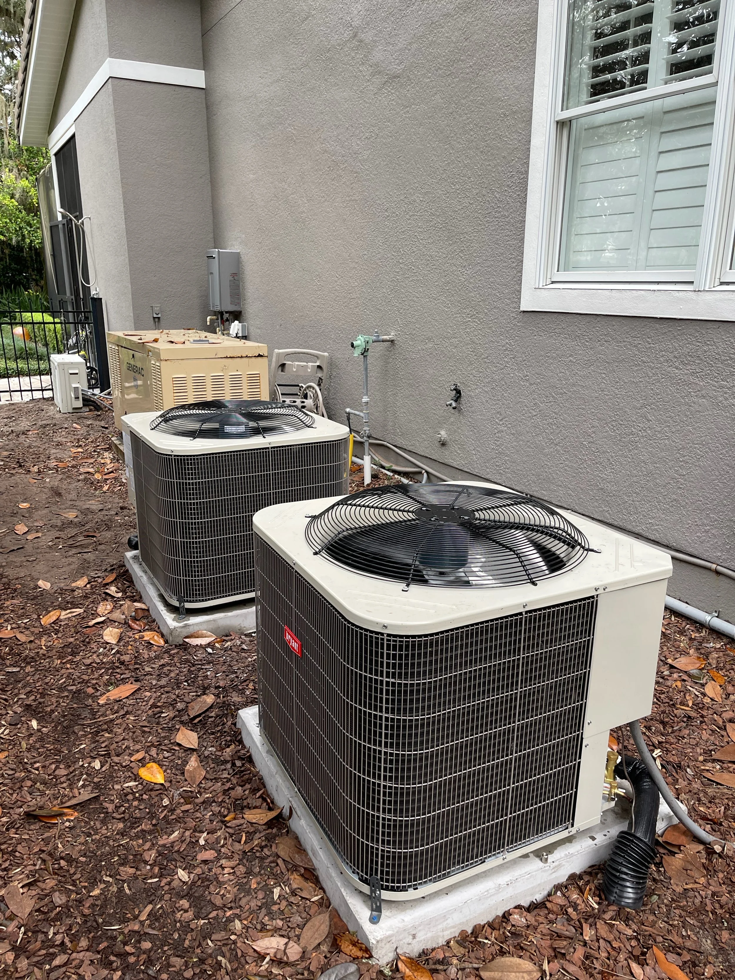 Outdoor view of three central air conditioning units installed on a concrete pad outside a house with a gray stucco exterior wall and a window with white shutters. Leaves are scattered on the ground, and pipes and electrical connections are visible.