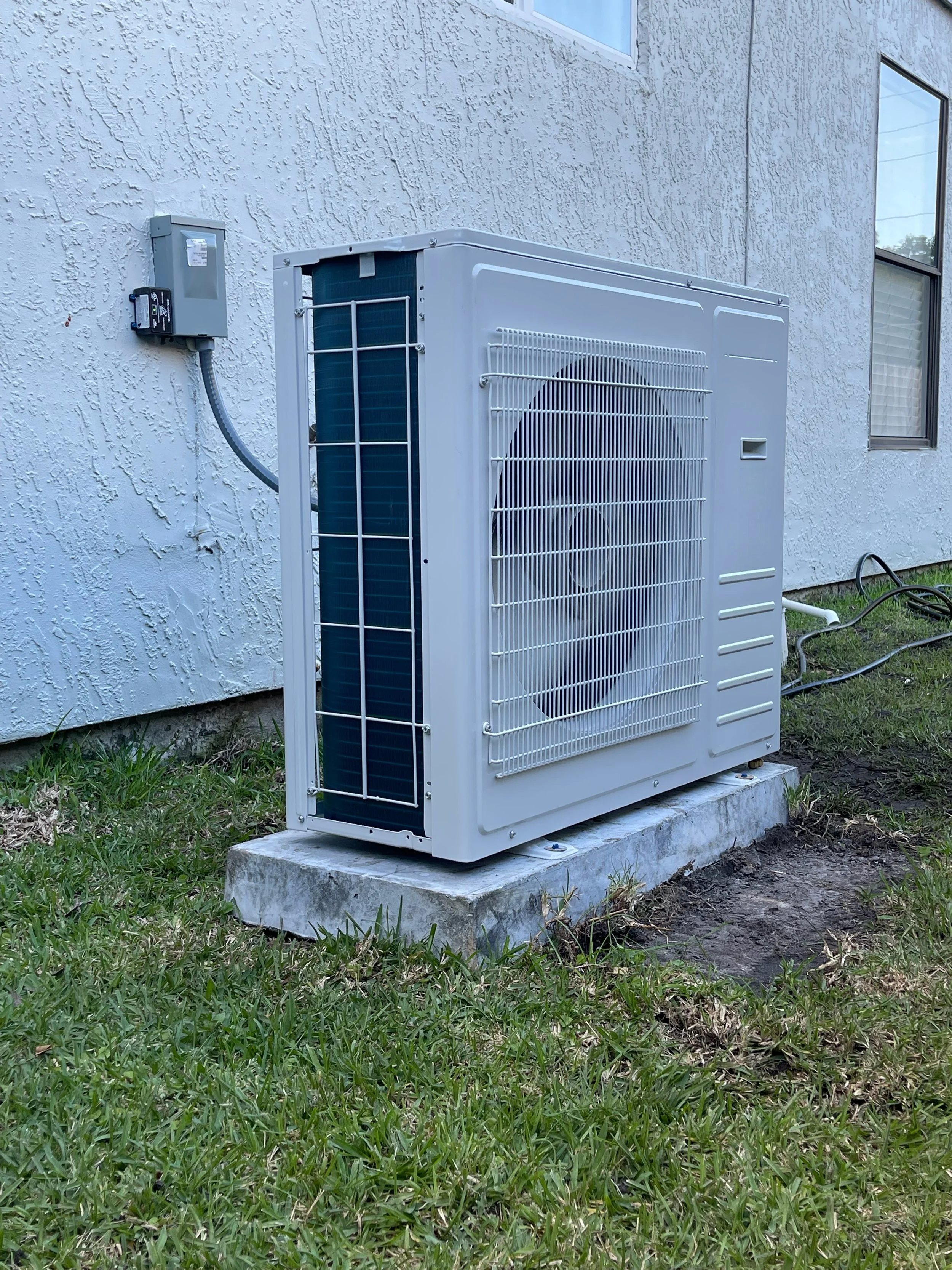Outdoor air conditioning unit installed on a concrete pad next to a textured white wall of a house.