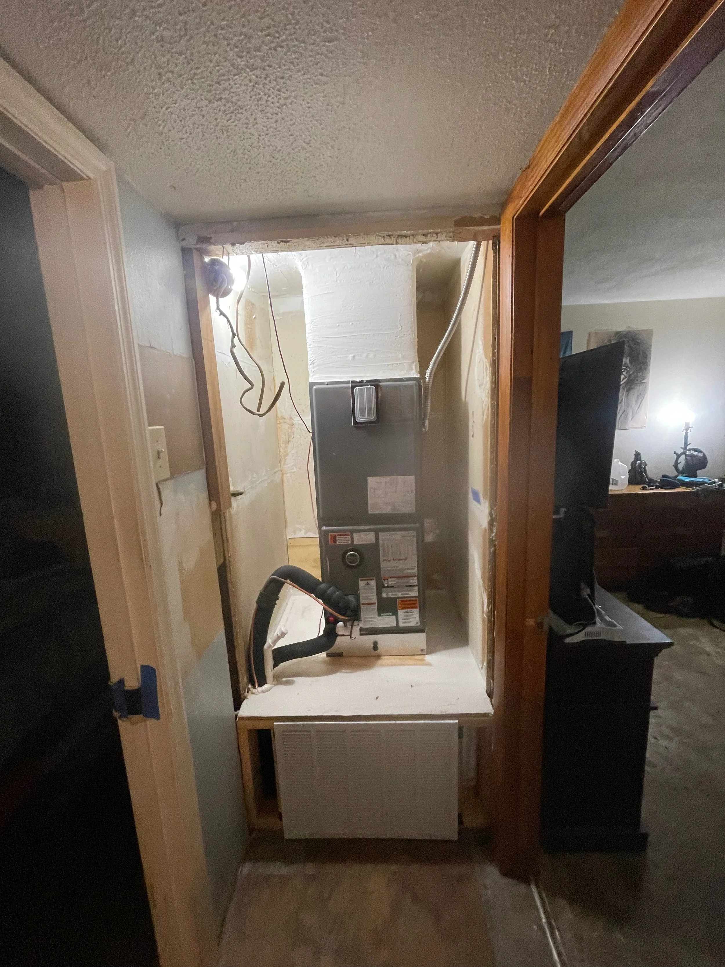 A small room with partially finished walls, containing a furnace or HVAC unit, with exposed wiring and pipes, and a wooden door frame.