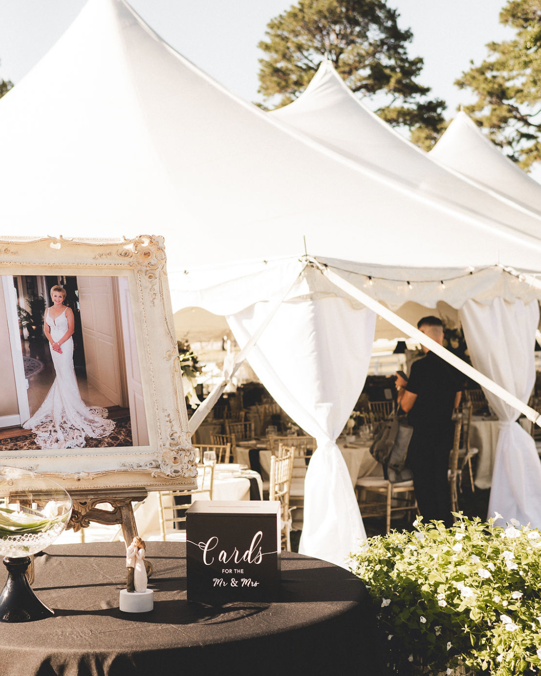Wedding reception area with a framed photo of a bride in a white gown, a black box labeled "Cards for the Mr. & Mrs," and a small angel figurine on a black table. White tents with open sides are in the background, with string lights and a person seen