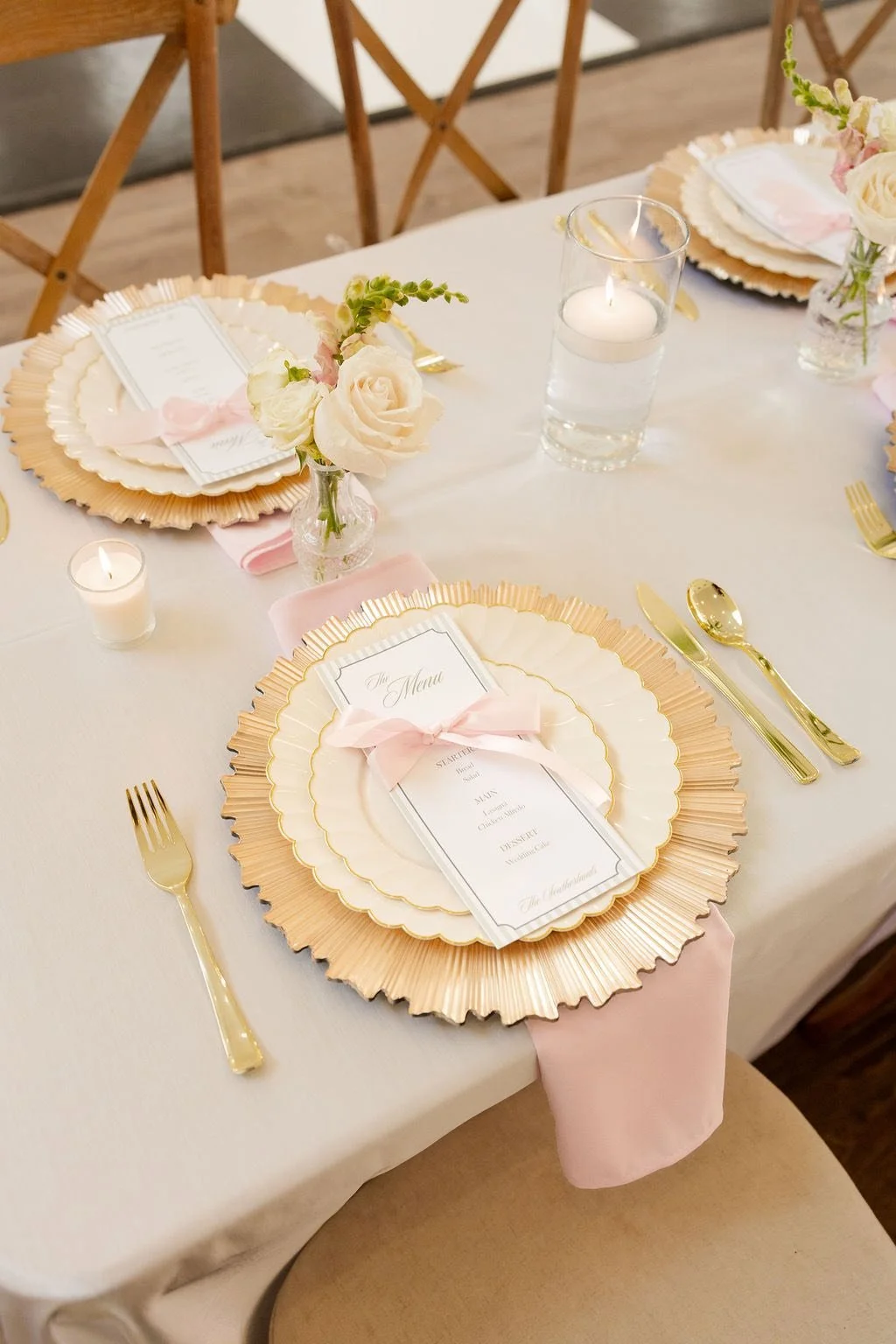 Elegant wedding table setting with gold and pink accents, featuring gold cutlery, a floral centerpiece with white roses and greenery, a candle in a glass holder, and a menu card tied with pink ribbon.
