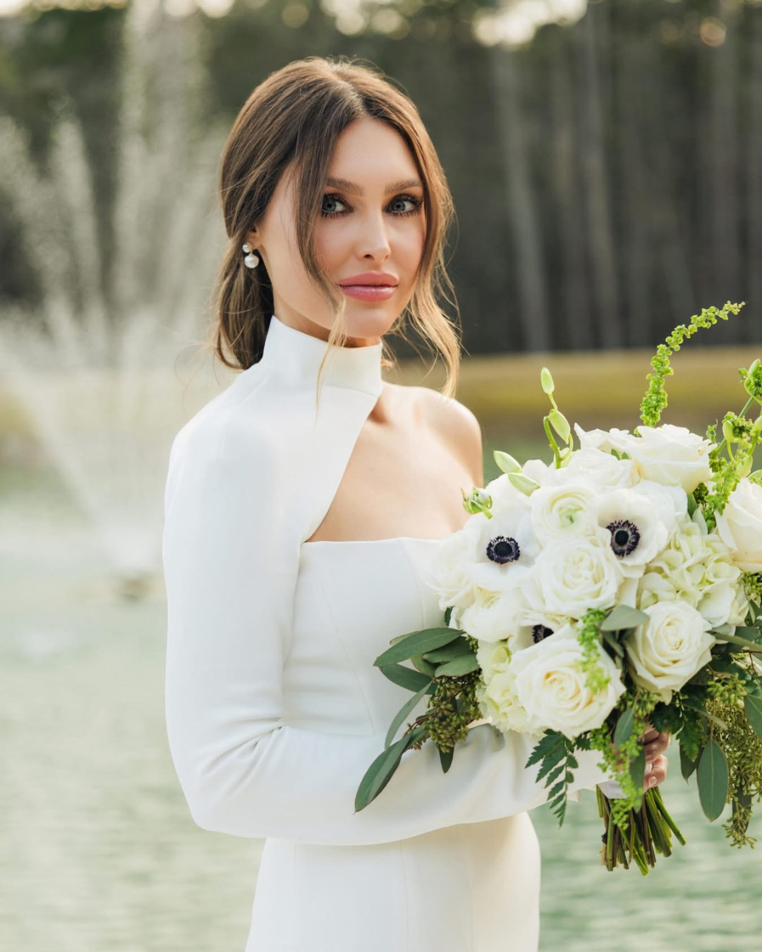 A bride holding a large bouquet of white roses and flowers outdoors, wearing a white dress with a high neckline, in front of a blurred natural background.